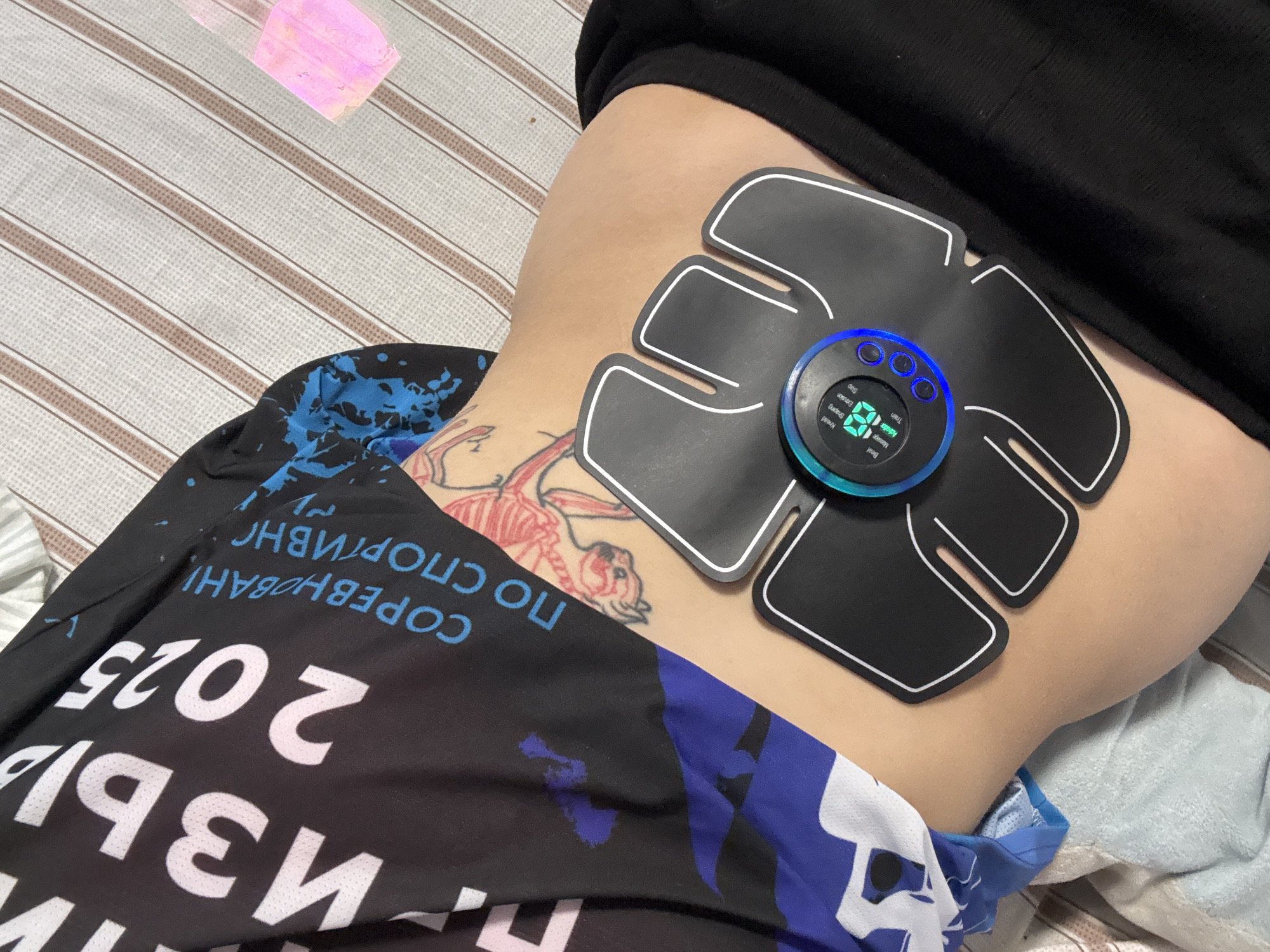 A person is lying on a striped surface. A black electrical muscle stimulation device is attached to their waist. The person is wearing a black shirt and athletic pants with writing in Russian and the year "2025". A small tattoo is visible on their skin. The setting appears to be an indoor environment. There is no definitive evidence in the image to establish a specific location in Прикамский, Russia.