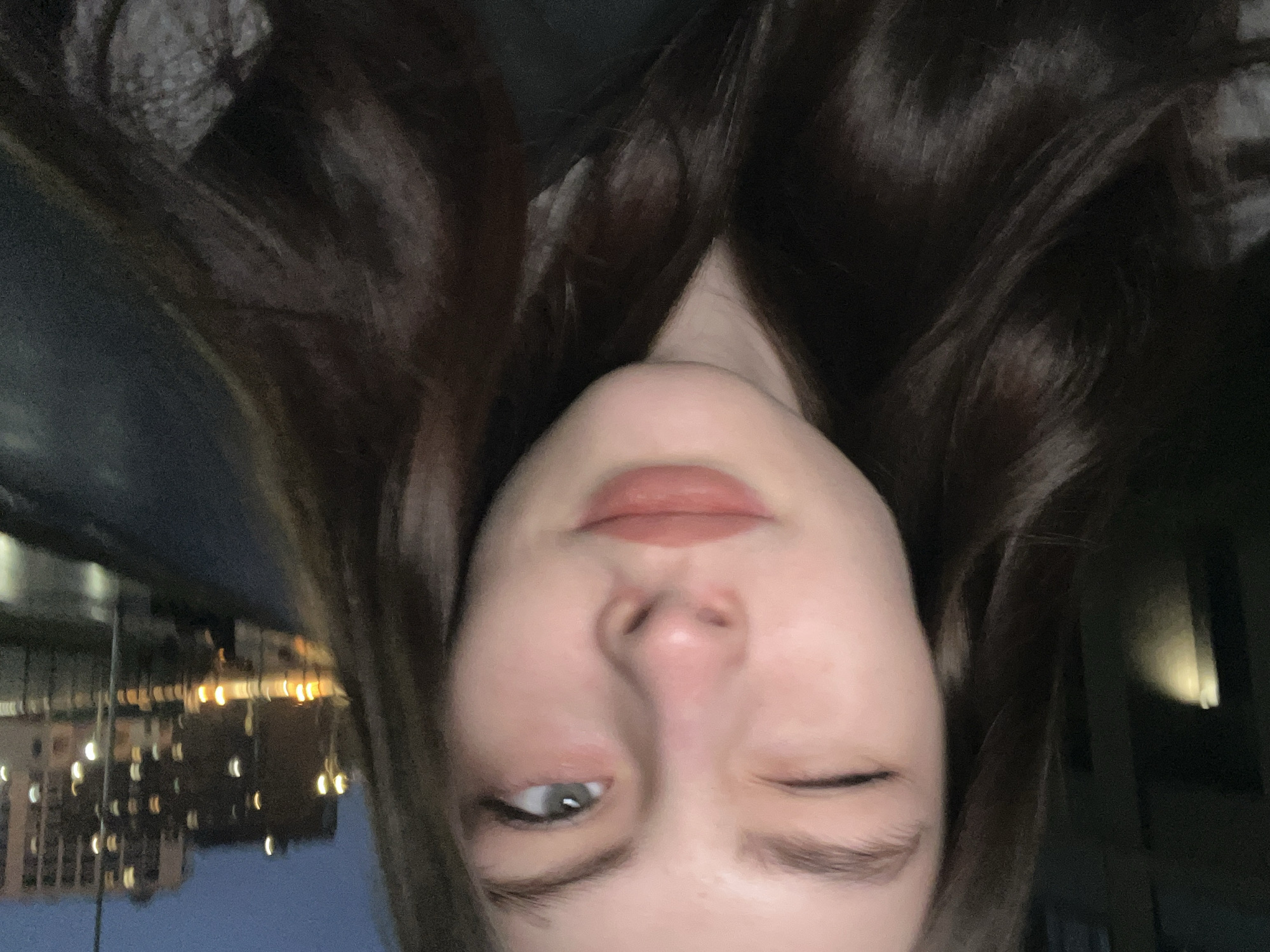 An upside-down selfie of a woman with long, dark brown hair and fair skin. She is winking with her right eye, and her left eye is visible and appears to be gray. She has on lipstick. Outside a window, building lights are visible, and the city is Saint Petersburg, Russia.