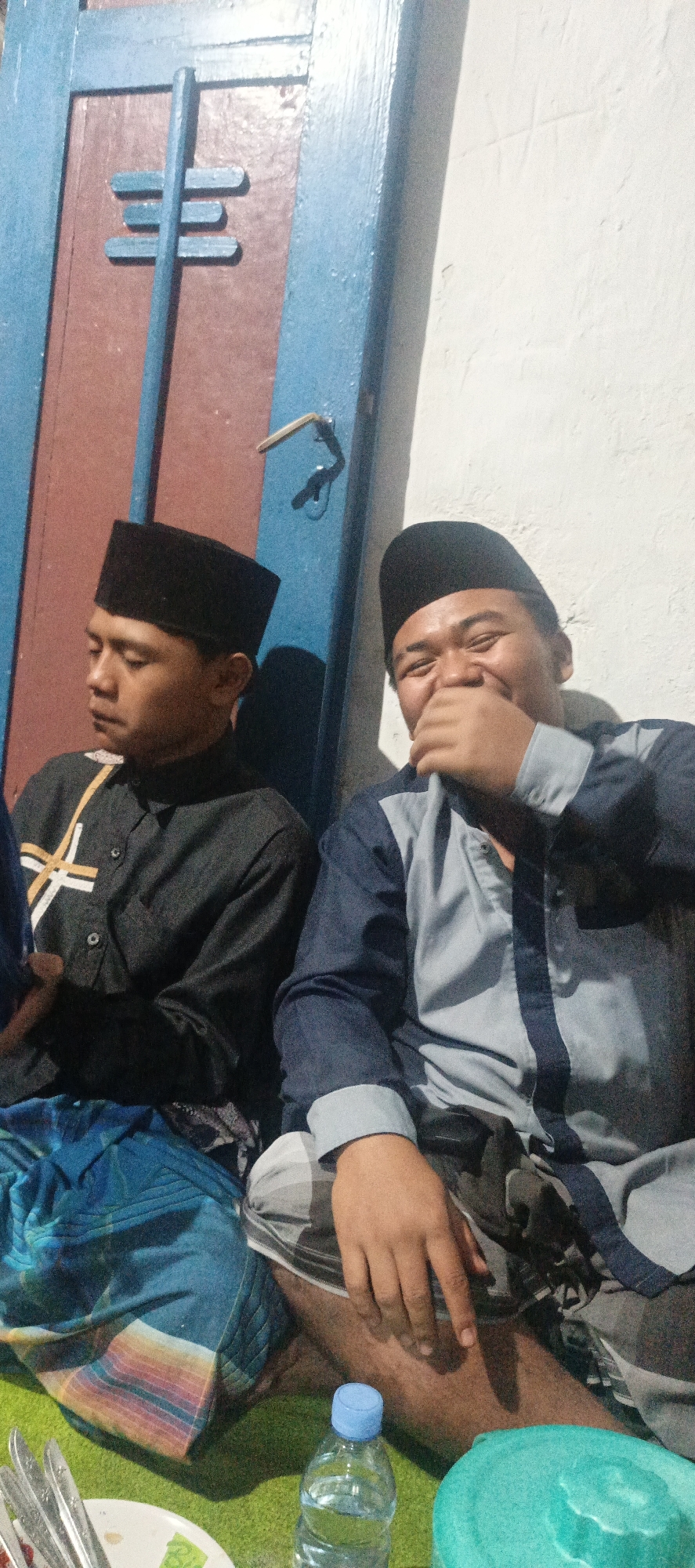 Two men are sitting next to each other in front of a blue and brown door. The man on the left is wearing a black peci and a black shirt with a yellow and white stripe design. He is looking down with his head tilted slightly. The man on the right is also wearing a black peci and a blue and grey collared shirt. He is laughing and covering his mouth with his hand. Both men are wearing patterned sarongs. The background is a textured white wall. In the foreground, there is a green mat, a plate with silverware, a bottle of water, and a turquoise object, possibly a bowl. The lighting is dim, casting shadows.