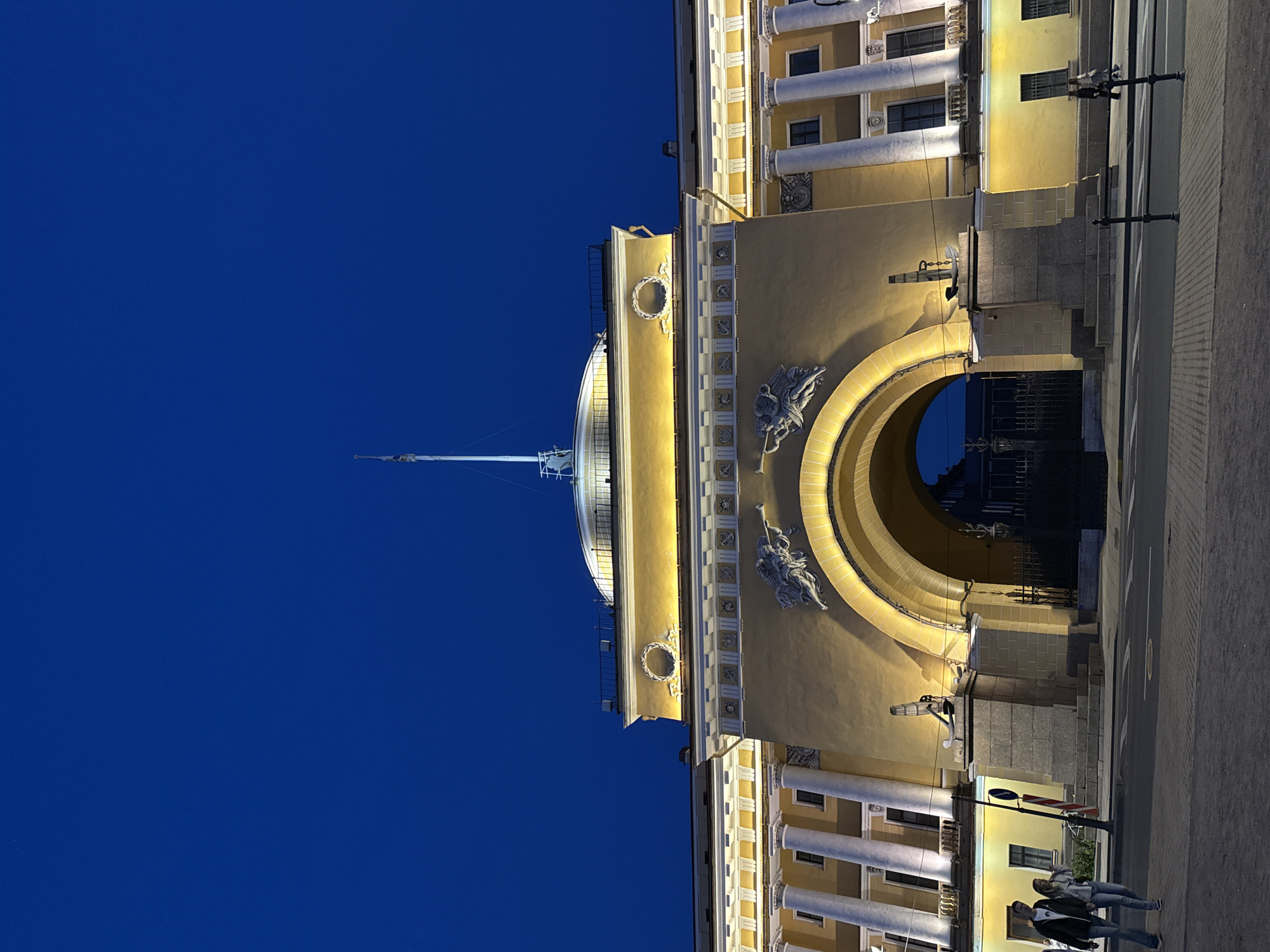 The image displays the grand facade of a monumental building, identified as the Admiralty in Saint Petersburg, Russia, captured during twilight or nighttime. The architecture is characterized by a warm, light yellow or ochre color, illuminated by artificial lights that highlight its classical details.
The central feature is a large, ornate archway glowing with a soft, warm light. Above and around this archway, sculptural elements are visible, including an angel-like figure holding a trumpet and a decorative wreath. The building features multiple levels with prominent white columns and dark-framed windows. Rising from a dome-like structure on the roof is a tall, slender flagpole or mast with visible rigging, piercing the deep, rich blue sky.
At street level, a paved area, likely a sidewalk or square, runs along the base of the building. Several street lamps are visible, casting light onto the ground and the lower sections of the structure. Two individuals are seen walking casually on the pavement towards the right side of the frame, dressed in everyday jackets and jeans.
The overall scene conveys a sense of architectural grandeur and historical significance, presented in the quiet beauty of an evening setting with distinct artificial illumination contrasting with the natural darkness of the sky. No discernible text is visible in the image.
