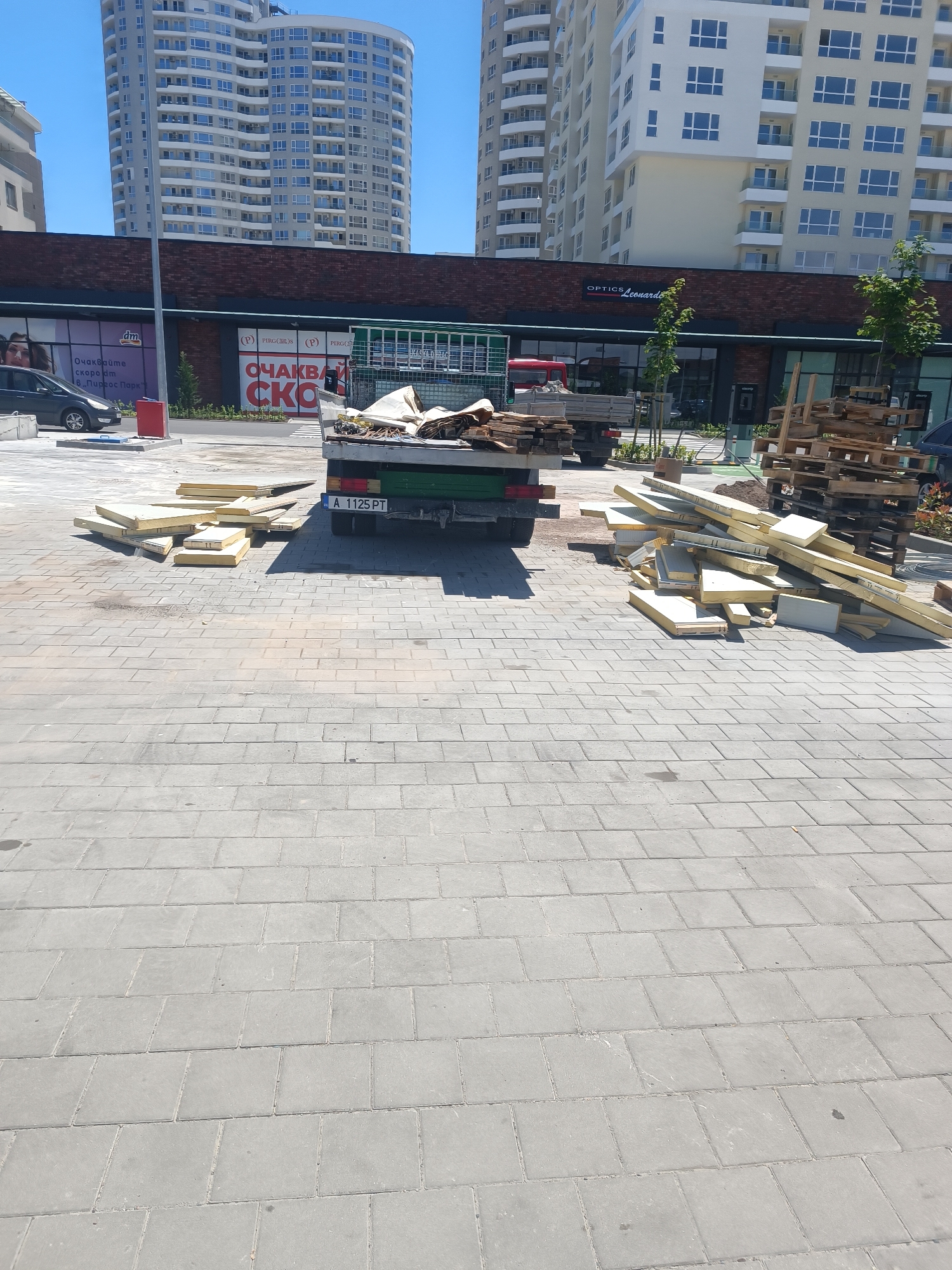 The image depicts a construction site located in the city of Burgas, Bulgaria. In the foreground, a truck bed is partially visible, loaded with what appears to be construction materials, possibly insulation panels and wooden planks. These materials have also been unloaded and are piled on the paved ground to the left and right of the truck. The truck itself has a license plate reading "A 1125 PT". The surrounding area is paved with gray rectangular blocks, characteristic of an urban walkway or plaza. In the background, a row of modern, multi-story buildings with numerous windows are visible, suggesting a commercial or residential complex. The sky is clear and blue, indicating a sunny day. There are no people clearly visible in the image. The overall scene conveys a sense of ongoing construction or renovation work in an urban environment.