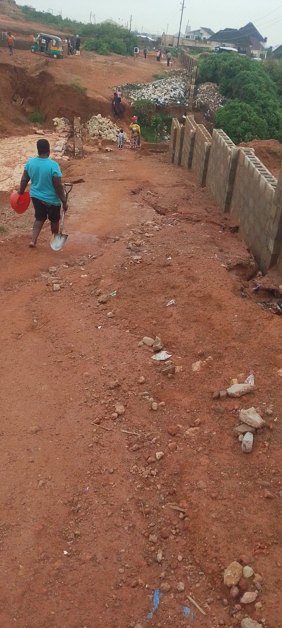 The image depicts a construction site in Rayfield, Nigeria, characterized by red dirt terrain and ongoing building activities. In the foreground, a person wearing a blue t-shirt and black shorts walks away from the camera, carrying a red bucket in their left hand and a shovel in their right. The ground is uneven and littered with small rocks and debris.
Further into the scene, a partially constructed brick wall runs along the right side of the frame, separating the construction area from an overgrown, vegetated embankment. Beyond the wall, several houses are visible on a hill in the background. In the middle ground, there is a significant pile of garbage situated on the embankment. Several individuals, including some children, are scattered throughout the site, with a tuk-tuk vehicle parked on a higher level of the terrain to the left. The overall atmosphere suggests a community undergoing development or repair, with evidence of both construction and waste management challenges.