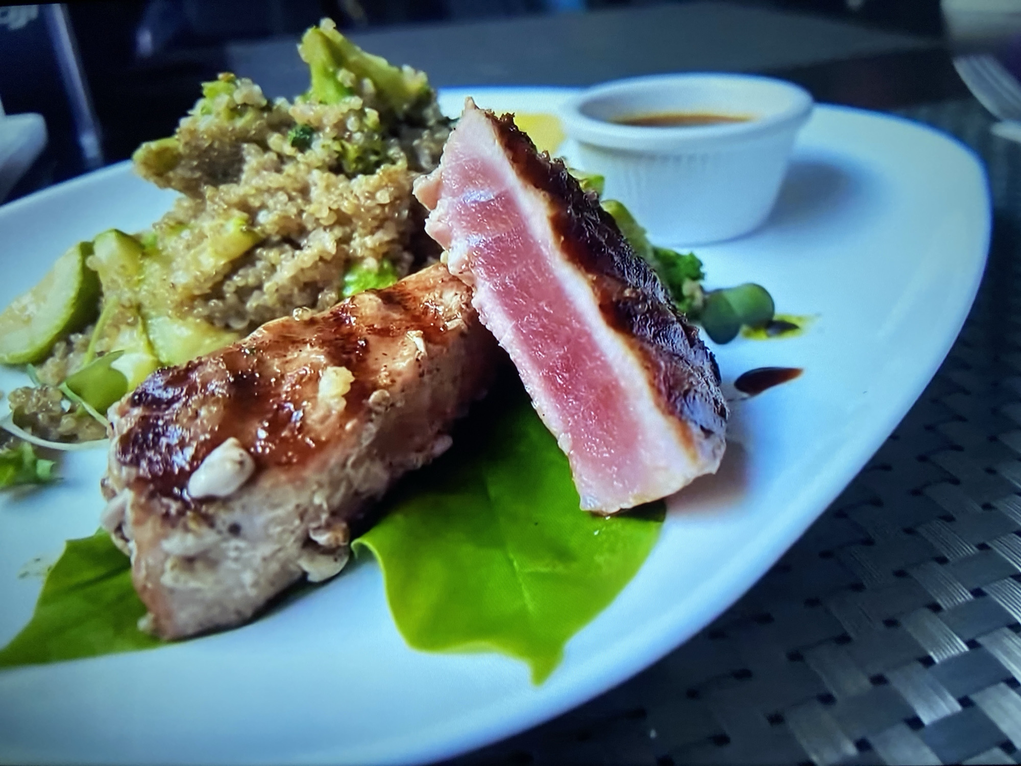 On a white plate with a slight blue hue, lies a culinary presentation consisting of seared tuna, a grain salad, and a small bowl of sauce. The tuna, browned on the outside and pink on the inside, is sliced and rests on a bright green leaf. Adjacent to the tuna is a mound of grain salad, possibly quinoa, mixed with green vegetables, possibly zucchini and broccoli. A small white bowl containing a brown sauce is positioned in the background. The plate rests on a woven surface, which appears to be a table. The scene suggests a dining establishment, potentially located in Unknown, Unknown, given the unknown location.