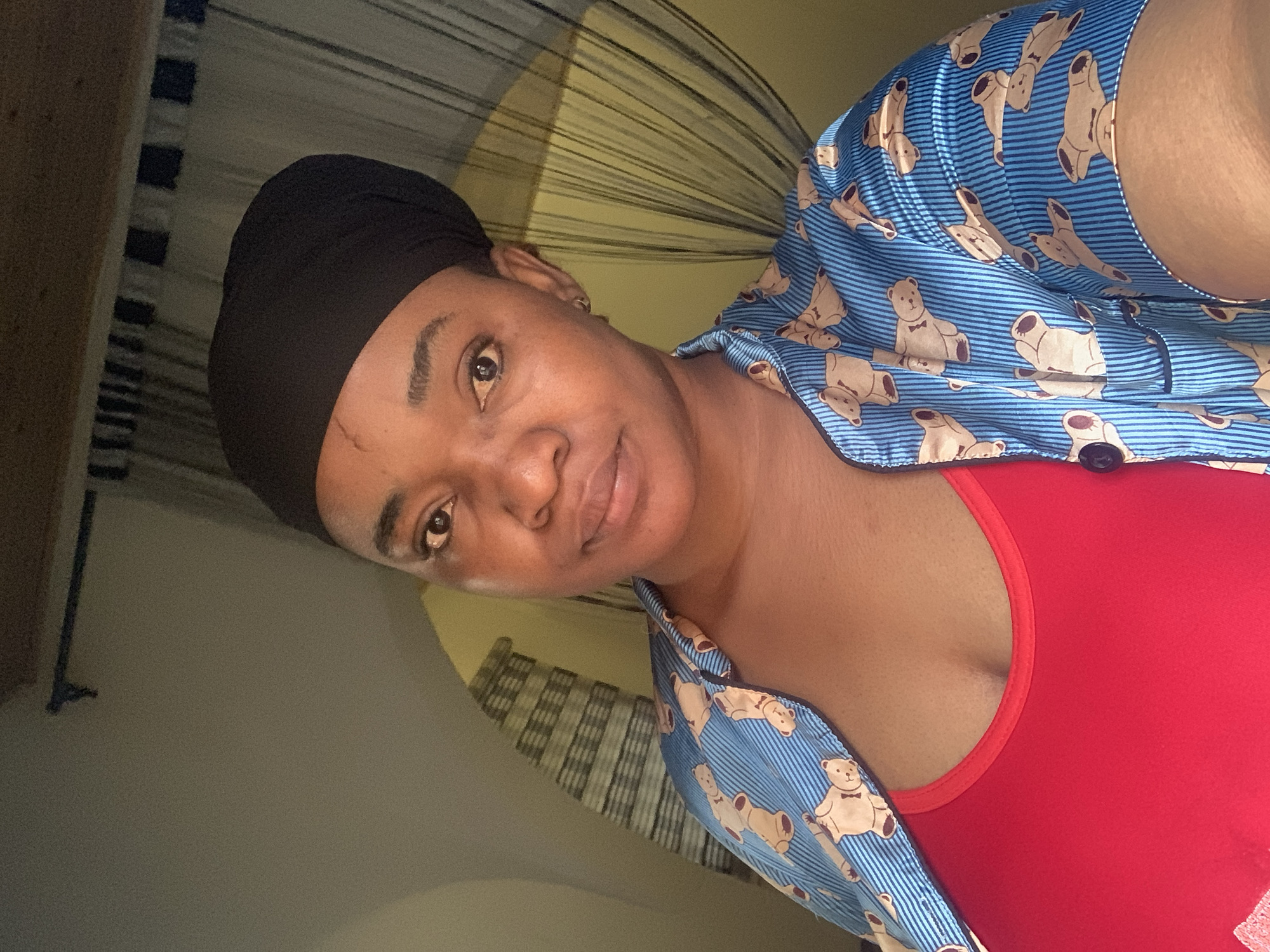 The image shows a woman wearing a black head covering, a red top, and a blue shirt with teddy bear patterns. The background includes a window covering and a wall. The image appears to be taken indoors. There is no information provided that indicates the image was taken in the city of Barakin Akawo, Nigeria.