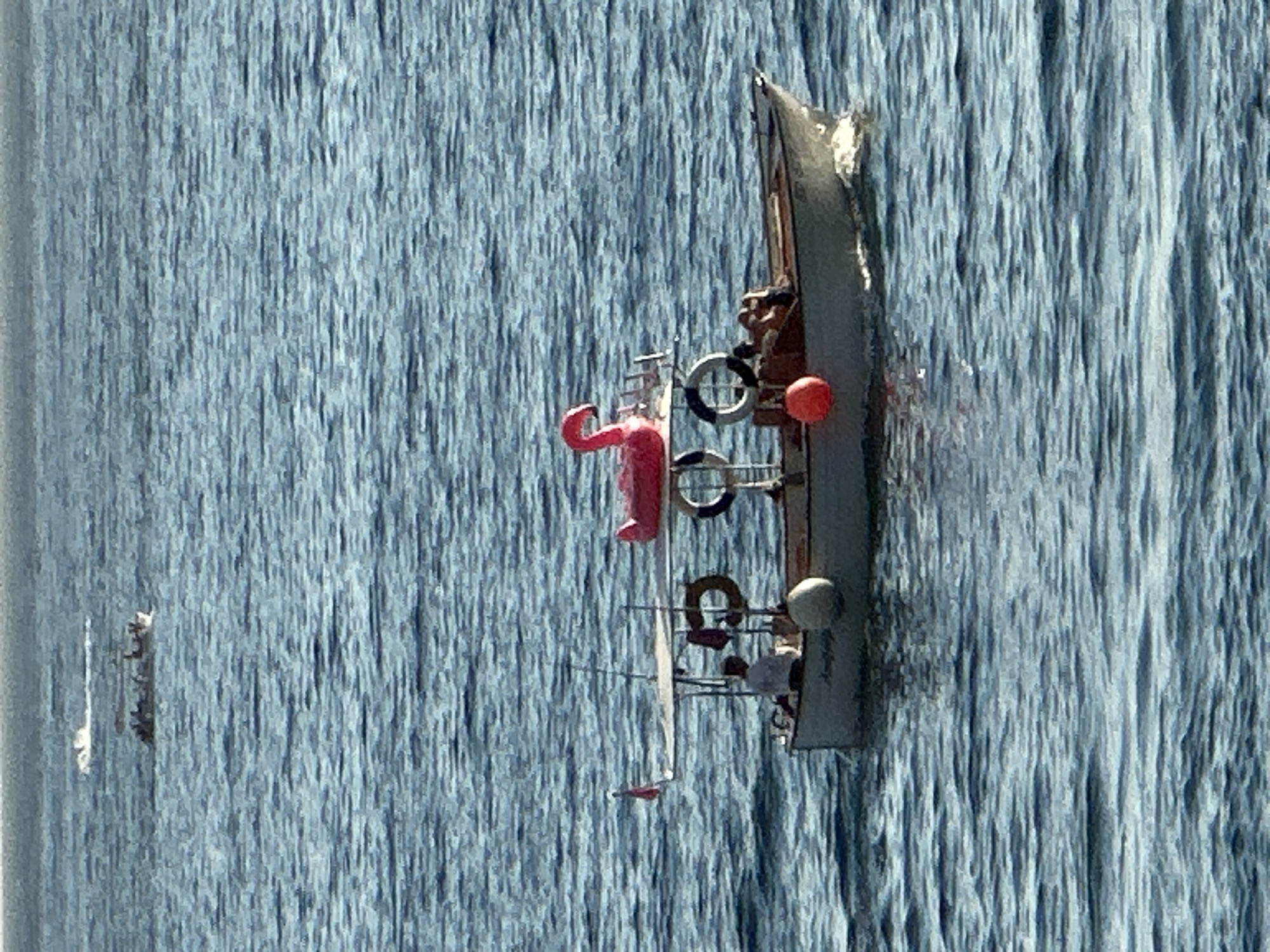 The image shows a body of water, likely the Adriatic Sea, with several boats visible. In the foreground, a small motorboat is moving from left to right. This boat has a canopy over the back section, where a person in a white shirt is seated. On the roof of the canopy, a pink inflatable flamingo is placed. There are life preservers and an orange buoy attached to the boat. Two people are visible in the stern of the boat, one appearing to be lying down. In the mid-ground, another boat, larger than the first, is carrying multiple passengers and is also moving. In the background, a third boat is visible as a small white object on the water, suggesting it is further away and moving at speed. The water is blue with visible ripples. The horizon line is visible in the upper part of the image. The scene appears to be a sunny day, based on the light and the calm water. Given the context of Budva, Montenegro, this scene is likely taking place in the waters off the coast of the city, possibly near the Budva Riviera or the Bay of Kotor.