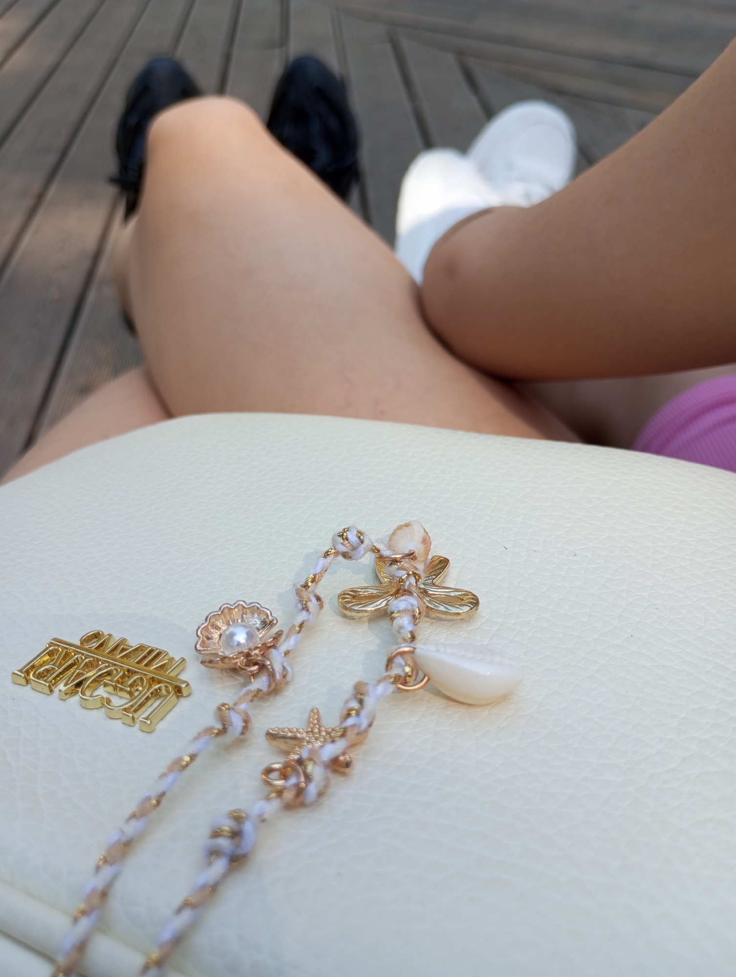 The image presents a first-person perspective of someone seated on a wooden surface, possibly a deck or boardwalk. The person is wearing black shoes on one foot and white sneakers on the other. In the foreground, on what appears to be a light-colored bag or surface, lies a necklace and a golden "Moschino Milano" brand badge. The necklace has a white and gold rope, and it has small shell and starfish charms. The background is blurry, but it appears to be an outdoor setting. There is no information available in this image to indicate that the location is Izhevsk, Russia.
