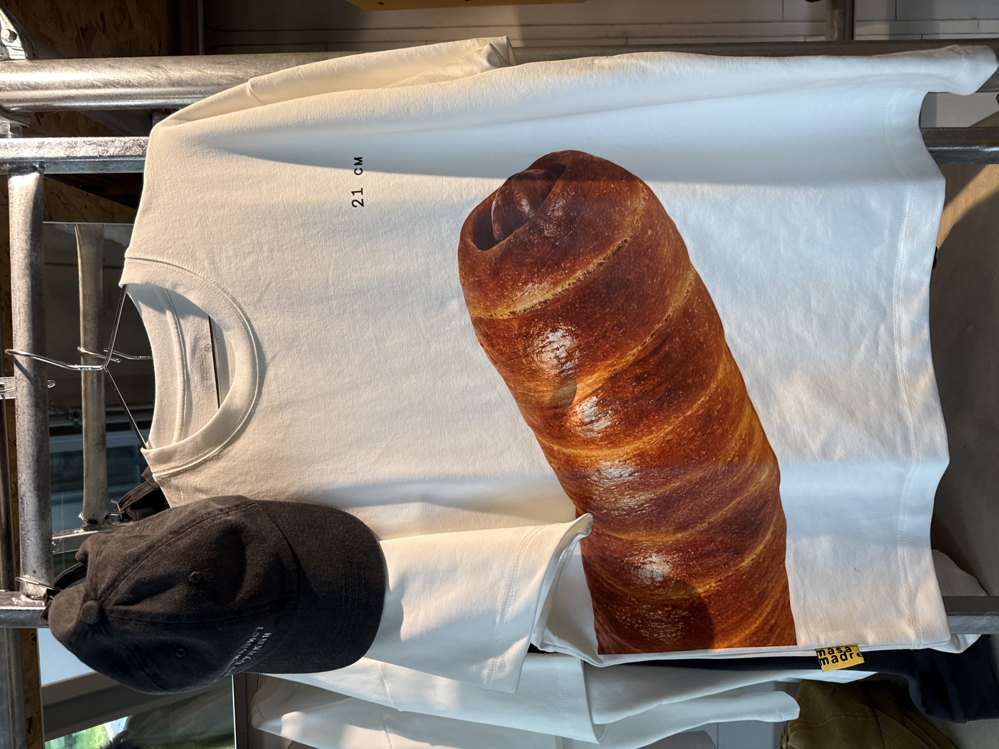 The image shows a white t-shirt with a realistic graphic print of a pastry, possibly a hot dog bun with a sausage inside, hanging on a metal rack. The t-shirt also has text on it, "21 CM" in black, located on the upper right side. Below the graphic, a yellow tag with black text that reads "masa madre" is visible. To the left of the t-shirt, a dark gray baseball cap is hanging, with some white text visible on its front panel. The background appears to be a retail environment, possibly a clothing store, with metal shelving and other garments. The overall lighting suggests an indoor setting. No people are visible in the image. The image does not provide any specific information about the location within the city of Moscow, Russia.