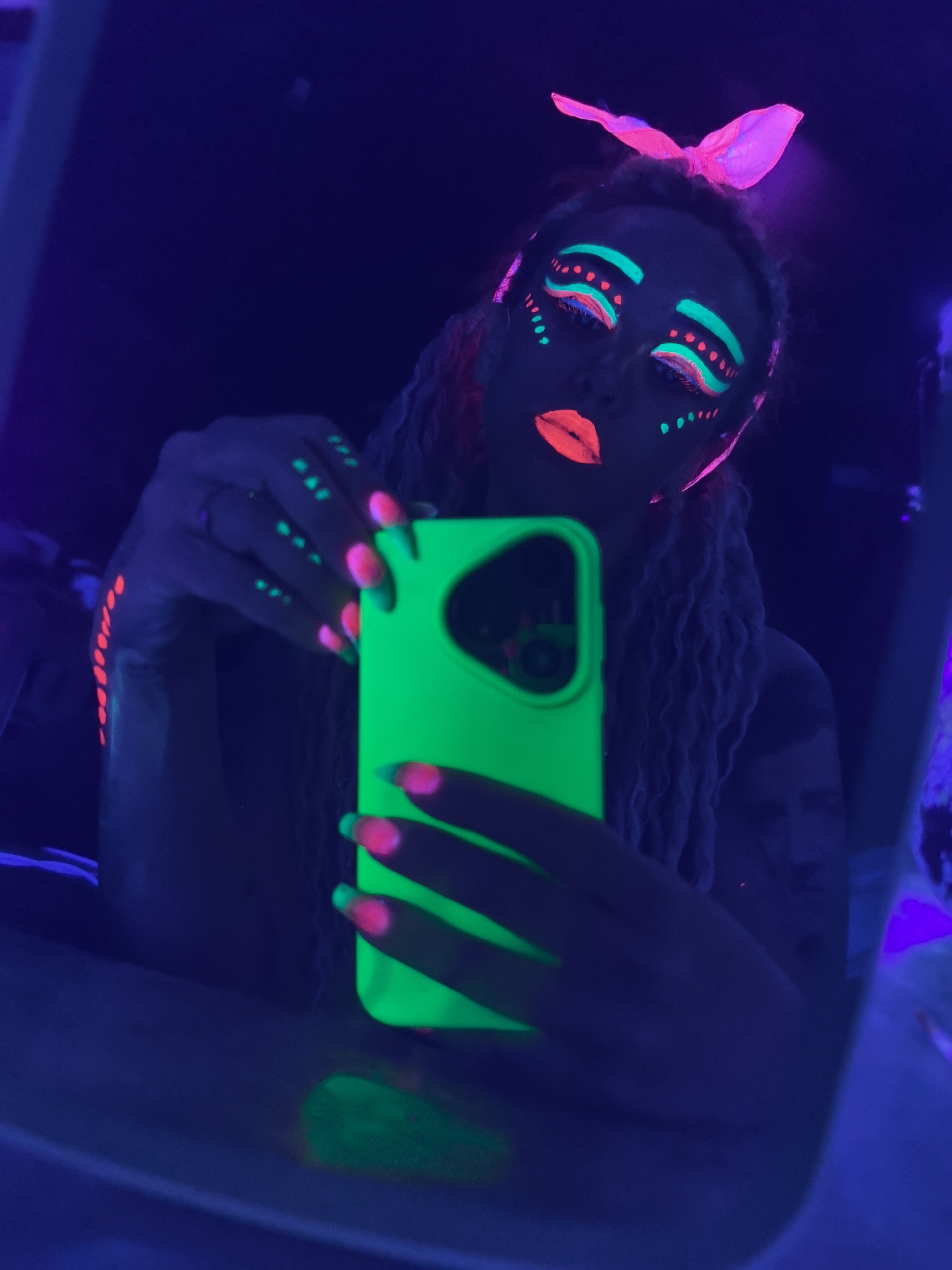 A person is taking a selfie in a dark environment illuminated by blacklights. The person has braids and wears a bright pink bow on top of their head. Their face is decorated with neon makeup: green arches above the eyes and dots near the eyes and cheeks, with red lipstick. Their fingernails are two-toned, with green and pink tips. They are holding a bright green smartphone. The background is dark and indistinct, suggesting an indoor setting with limited light sources other than the blacklights. The image appears to be a reflection in a mirror. There is no indication of the location being in Noviy, Russia.