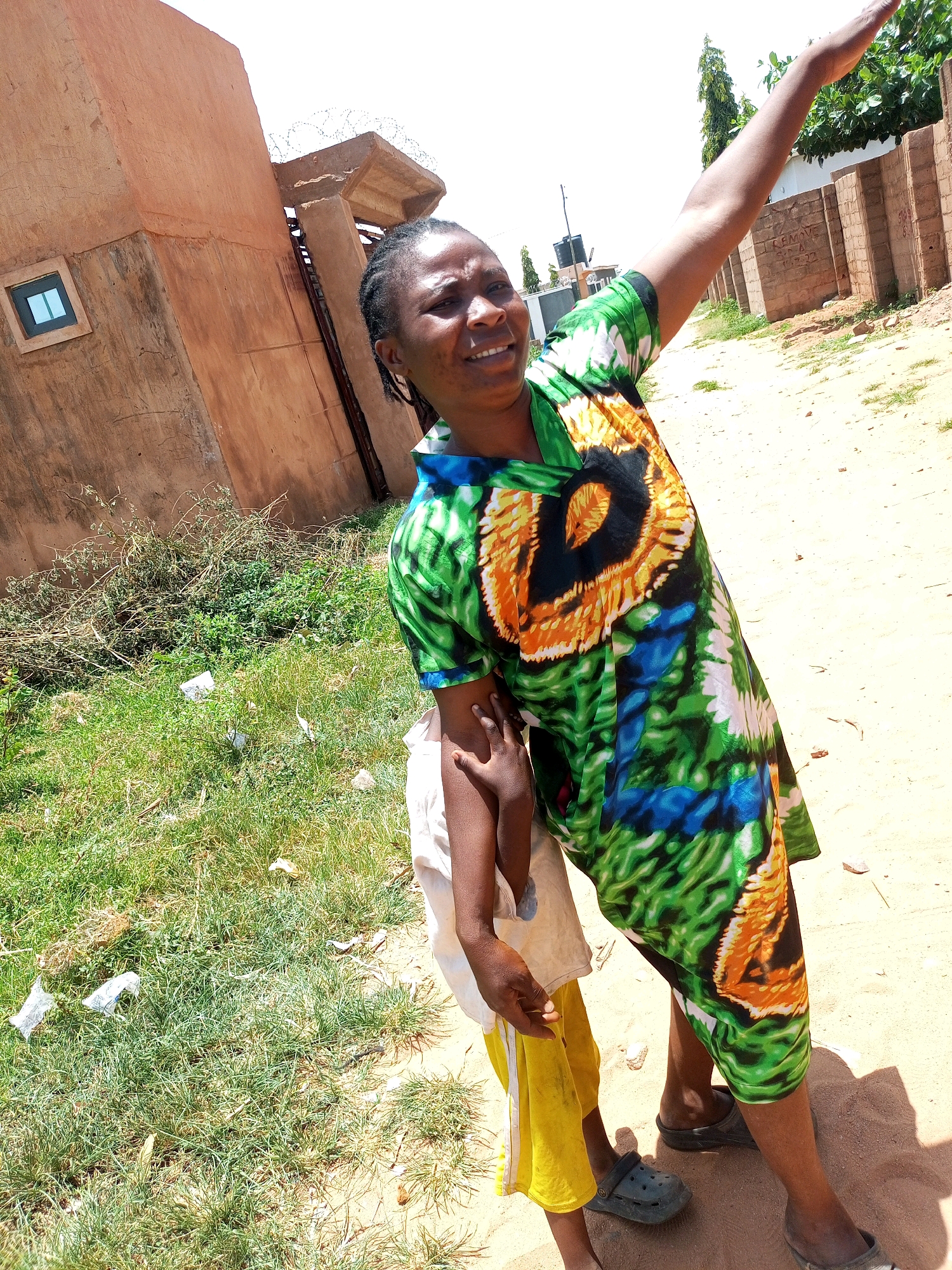 A Nigerian woman wearing a colorful African print dress is standing next to a young child in yellow pants and a white shirt. The woman is raising her right arm and pointing upwards. The child is holding onto the woman's arm. They are standing on a dirt path next to a grassy area with some litter. In the background, there is a beige building with a small window, a wall with barbed wire, and a brick wall. The scene appears to be outdoors in an urban or semi-urban area.