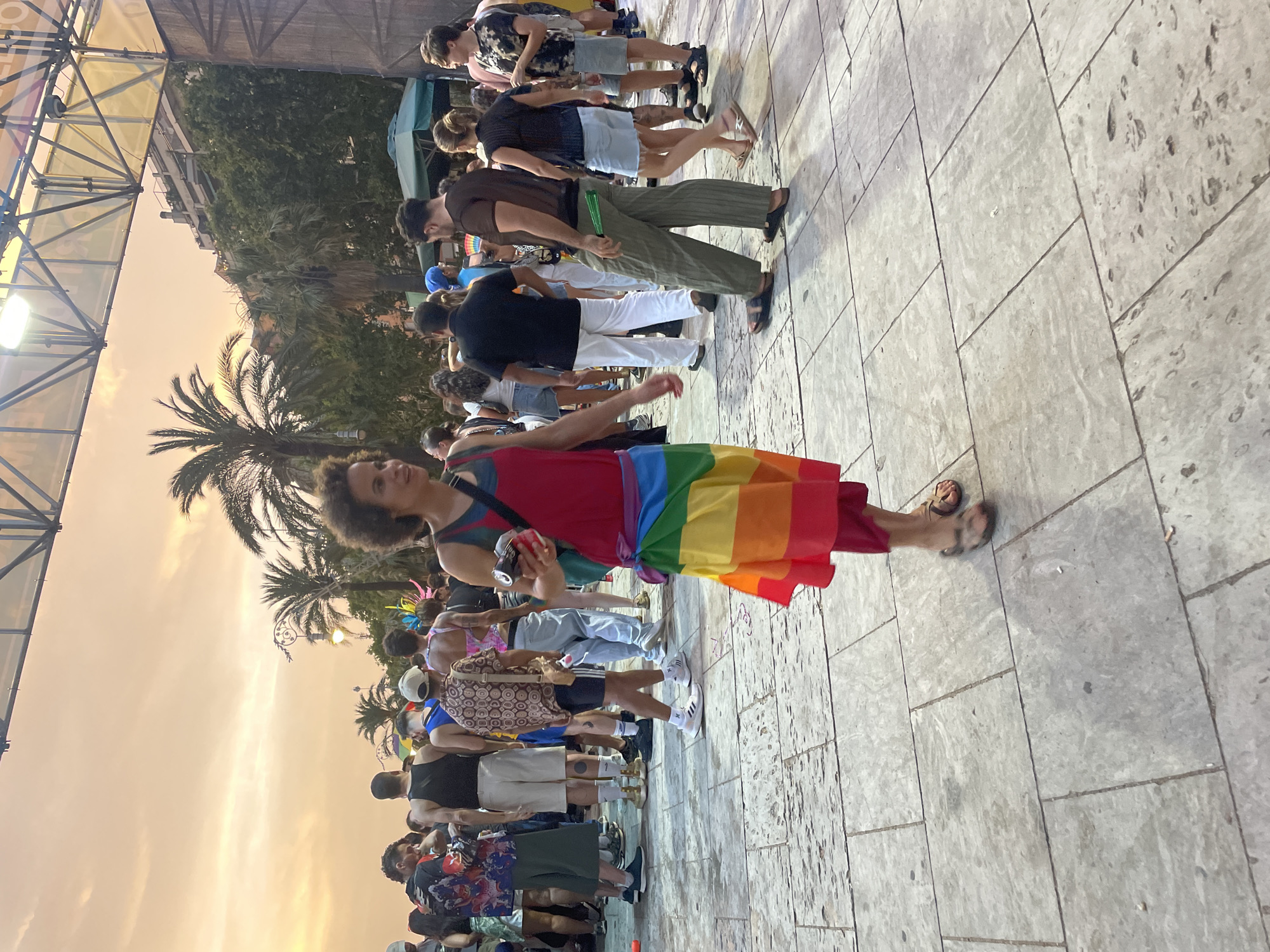 A group of people are gathered in what appears to be a city square in Barcelona, Spain, during a pride event. The sky is a warm orange hue, suggesting it is either sunrise or sunset, with palm trees visible in the background. The ground is paved with large, grey stone tiles. In the foreground, a person wearing a vibrant rainbow-colored dress is walking towards the camera, holding a can in their right hand. They have curly hair and are wearing sandals. Other individuals in the crowd are dressed in casual summer attire, with some wearing t-shirts, shorts, and other colorful clothing. There is a stage structure with lights visible in the background, indicating a performance or event is taking place. The overall atmosphere seems festive and celebratory.