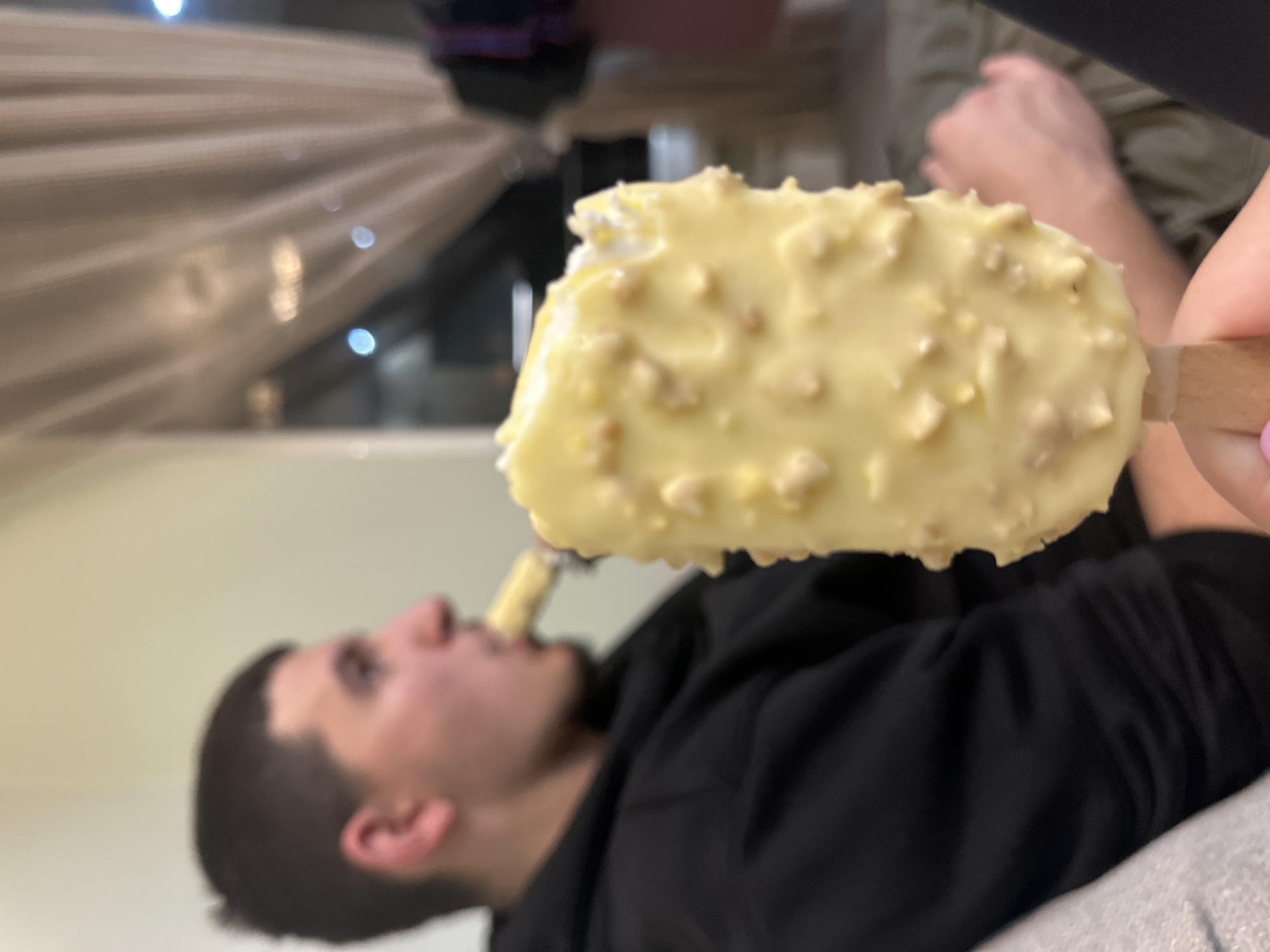 The image depicts an indoor scene with a focus on two individuals consuming ice cream bars. In the immediate foreground, a hand, exhibiting a pink-painted fingernail, holds a light-yellow, nut-coated ice cream bar on a wooden stick; a portion is missing from its upper right. In the midground, to the left, a blurry male subject with short, dark hair, wearing a black hoodie, is visible in profile, actively consuming a similar ice cream bar. The background consists of a light-colored wall, obscured furniture, and a window covered by light-colored, pleated curtains through which blurry artificial lights are visible. Indistinct parts of a third individual are discernible in the far right background. The specific location within Sofia, Bulgaria, cannot be determined from the visual information provided.