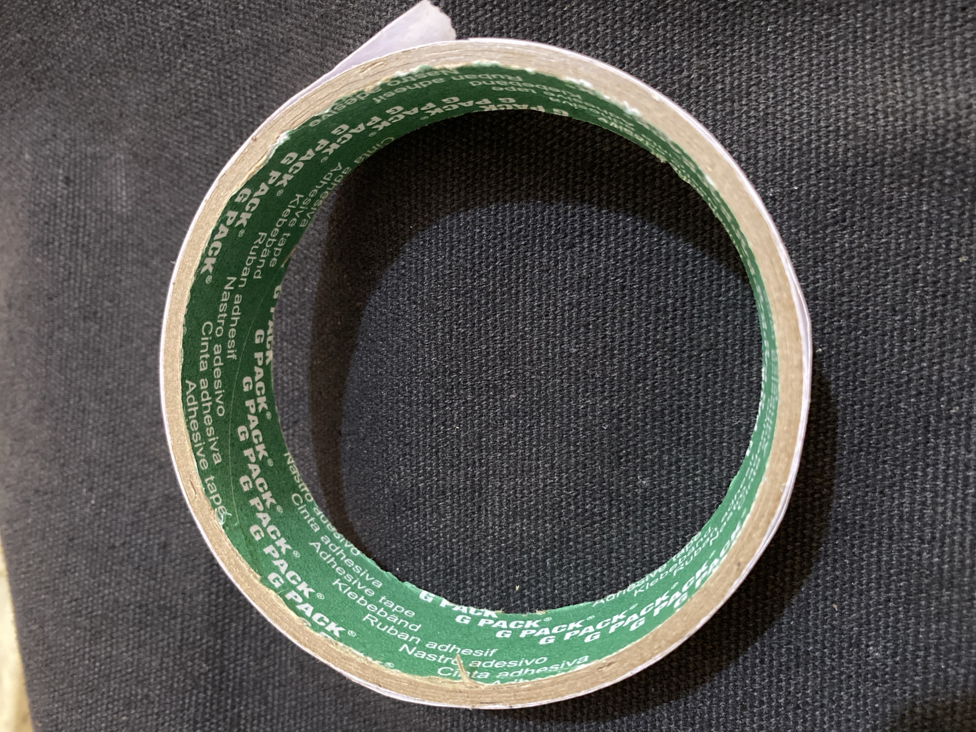 A roll of double-sided adhesive tape is placed on a dark, textured surface. The tape has green paper backing with repeating white text that reads "G PACK" along with various other languages like "Nastro adhesif", "Cinta adhesiva", "Klebeband", and "Adhesive tape". The tape is coiled into a circle, showcasing its construction. The background is a dark, woven fabric, possibly a textile or carpet. The lighting suggests an indoor setting, casting a subtle shadow on the surface. There are no people present in the image, and no discernible elements place the scene specifically within Lhokseumawe, Indonesia.