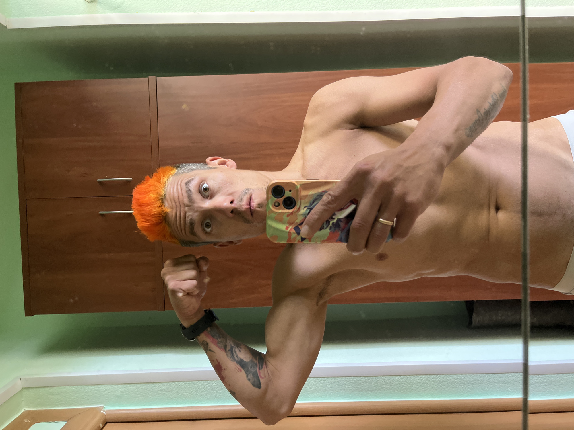A shirtless man with orange hair takes a selfie in a bathroom in front of a mirror. The man is flexing his right arm, which has a tattoo, and holding a phone with a patterned case in his left hand. He is wearing a wedding ring on his left ring finger. Behind him are brown cabinets and light green walls. The location appears to be a domestic bathroom. I cannot confirm the image was taken in Chaikovsky, Russia.