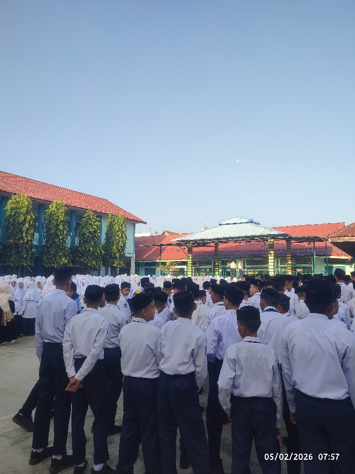 A large group of students in matching uniforms are standing in a schoolyard. They wear white shirts and dark pants. In the background, there are buildings with red tile roofs and green trim. The students appear to be gathered for an event or ceremony. A few people can be seen standing on a raised platform or stage. The sky is clear and blue. The date 05/02/2026 and the time 07:57 are visible in the bottom right corner.