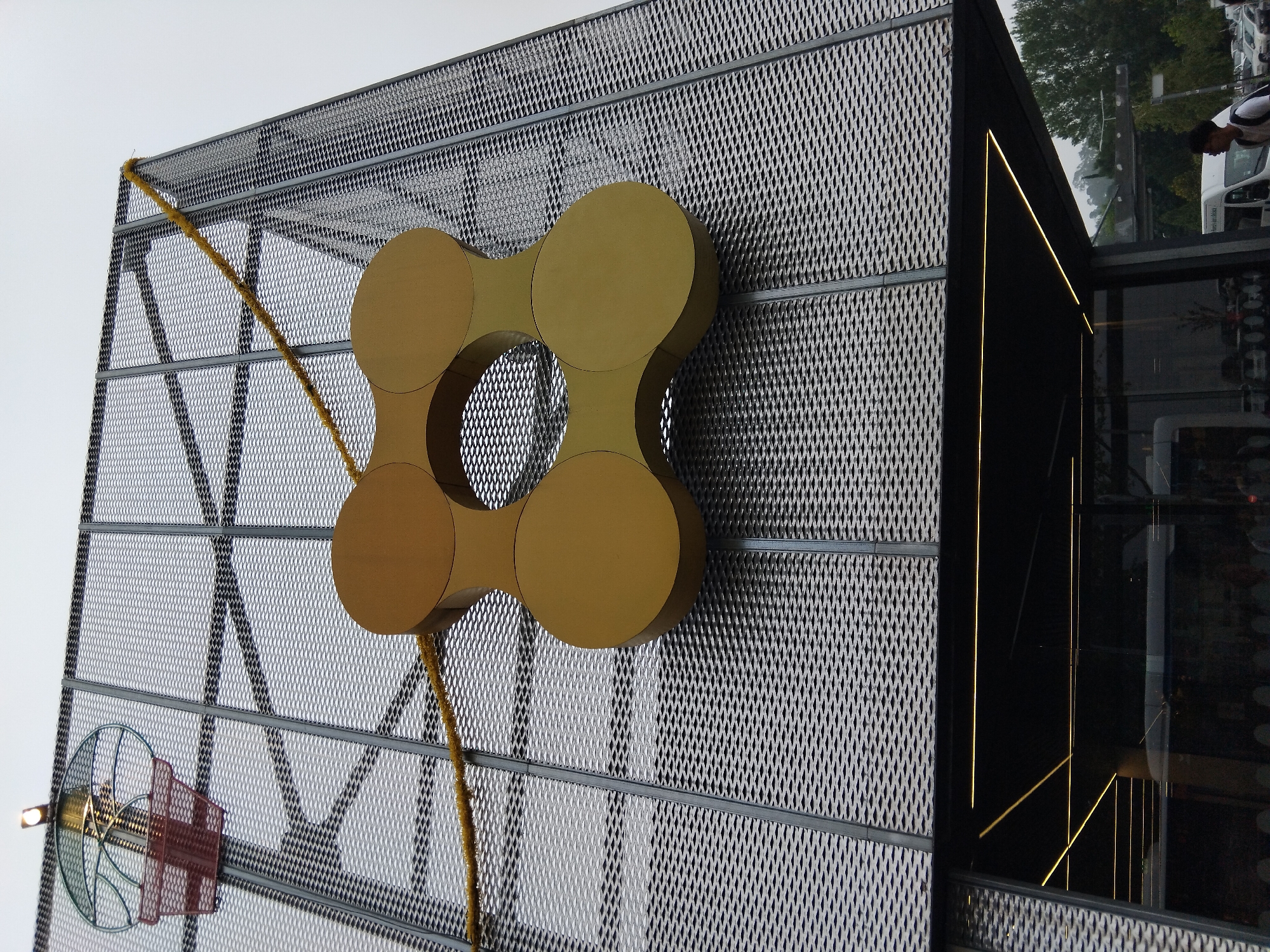 A low-angle, eye-level shot captures the facade of a modern building in Vila Nova de Famalicão, Portugal. The building features a large, metallic, diamond-patterned mesh facade. Mounted prominently on this mesh is a large, golden, four-lobed circular sculpture. A yellow, textured garland runs diagonally across the upper portion of the mesh. Below the mesh facade, a black overhang with embedded yellow lights is visible. Through the glass beneath the overhang, interior lights and reflections of people and vehicles can be seen. To the right, a person wearing a backpack walks past a white van. Trees and a cloudy sky are visible in the background.