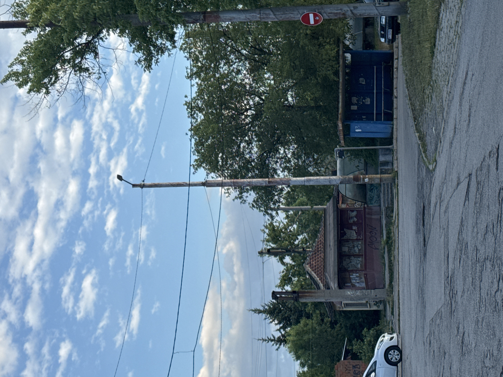 This is an outdoor, vertical, eye-level medium shot of a street scene in Pernik, Bulgaria. The sky is blue with scattered white clouds, suggesting it is daytime with fair weather. The scene includes a paved road, a grassy area, trees, overhead wires, and several structures that appear to be abandoned or under construction.
Prominent objects in the frame include a large blue dumpster, a small, white car parked on the right side of the road, and a dilapidated building with a red tiled roof. There are also utility poles and what looks like a traffic sign with a red circle and a white bar, indicating a "no entry" or similar restriction. Graffiti is visible on some of the structures, adding to the sense of neglect. The overall impression is of an urban fringe or an industrial area that is not well-maintained. The lighting suggests it is likely late afternoon, given the angle of the sun casting shadows. No people are visible in the image.