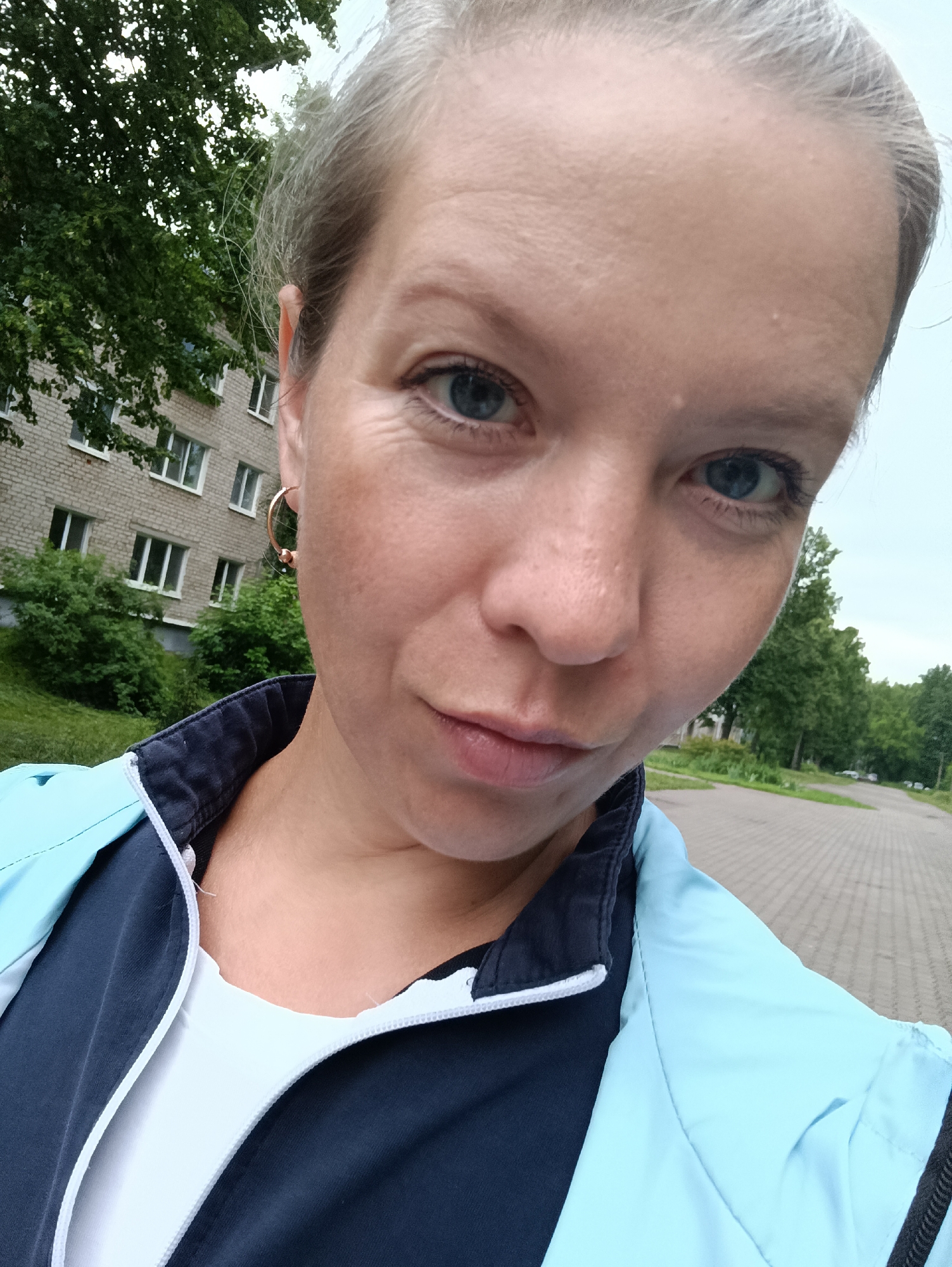 A close-up, high-angle selfie shows a fair-skinned woman with blonde hair pulled back, blue eyes, and light makeup. She is wearing a light blue and navy blue jacket over a white t-shirt. She has a gold hoop earring in her left ear. The background features a brick apartment building with green trees and bushes in front of it. To the right, a paved walkway with a slight curve stretches into the distance, lined with trees. The sky is overcast. This appears to be a residential area in Chaikovsky, Russia.