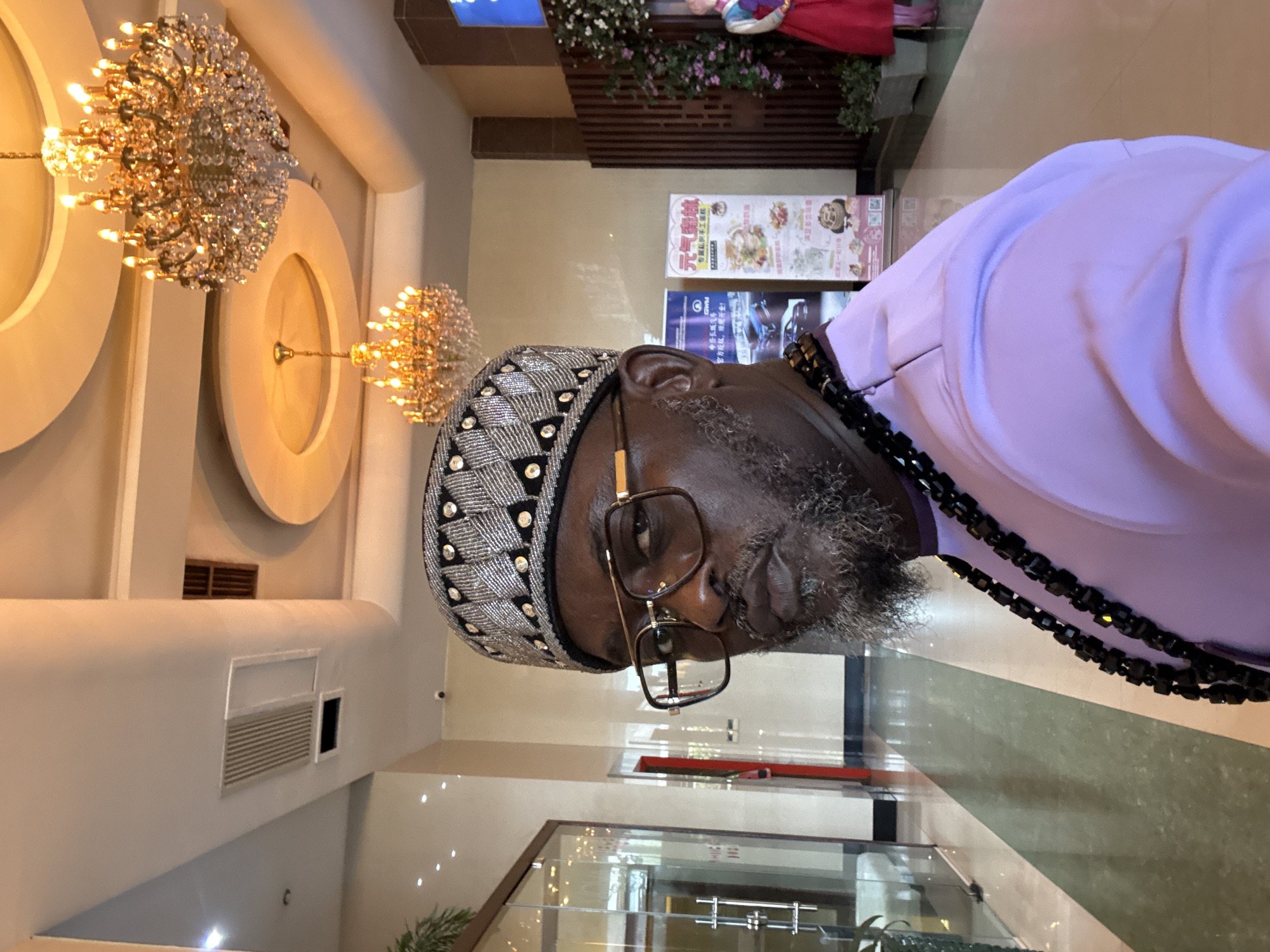 A middle-aged man with a grey beard, wearing glasses and a patterned hat, stands in a well-lit interior space. The man is dressed in a light purple shirt and a black beaded necklace. Behind him, ornate chandeliers hang from the ceiling, and there are circular design elements on the wall. The man is standing near a glass wall with greenery visible behind it. Further back, there are advertisements displayed on a wall. There is no indication of the city of Itirin, Nigeria.