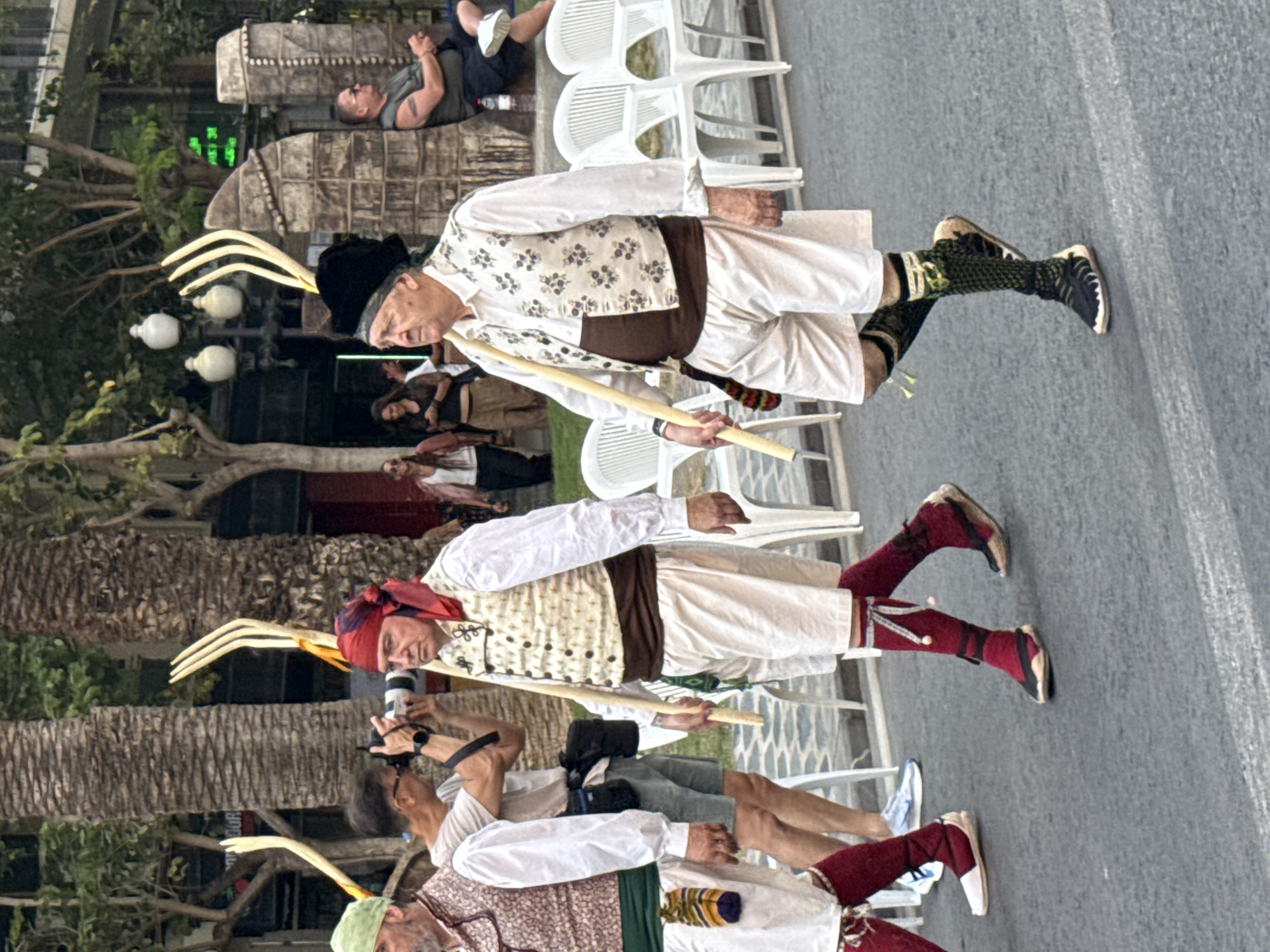The image captures a street scene in a city. Two men in traditional attire are walking forward, their faces turned to the left. The man on the left wears a red headscarf, a white vest with decorative patterns over a white shirt, white shorts, and red socks with white trim. He carries a wooden stick with a three-pronged fork at the top. The man on the right wears a black hat, a white vest with floral patterns over a white shirt, white shorts, and patterned green and black socks. He also carries a similar wooden stick.
In the background, several palm trees are visible, suggesting a warm climate. A person in the middle ground is taking a photograph with a large camera. Other individuals are present in the background, some sitting on white chairs, and others standing. A stone archway structure is also visible on the right side of the image. The ground is paved asphalt with white lines. The overall impression is of a cultural event or festival taking place outdoors.