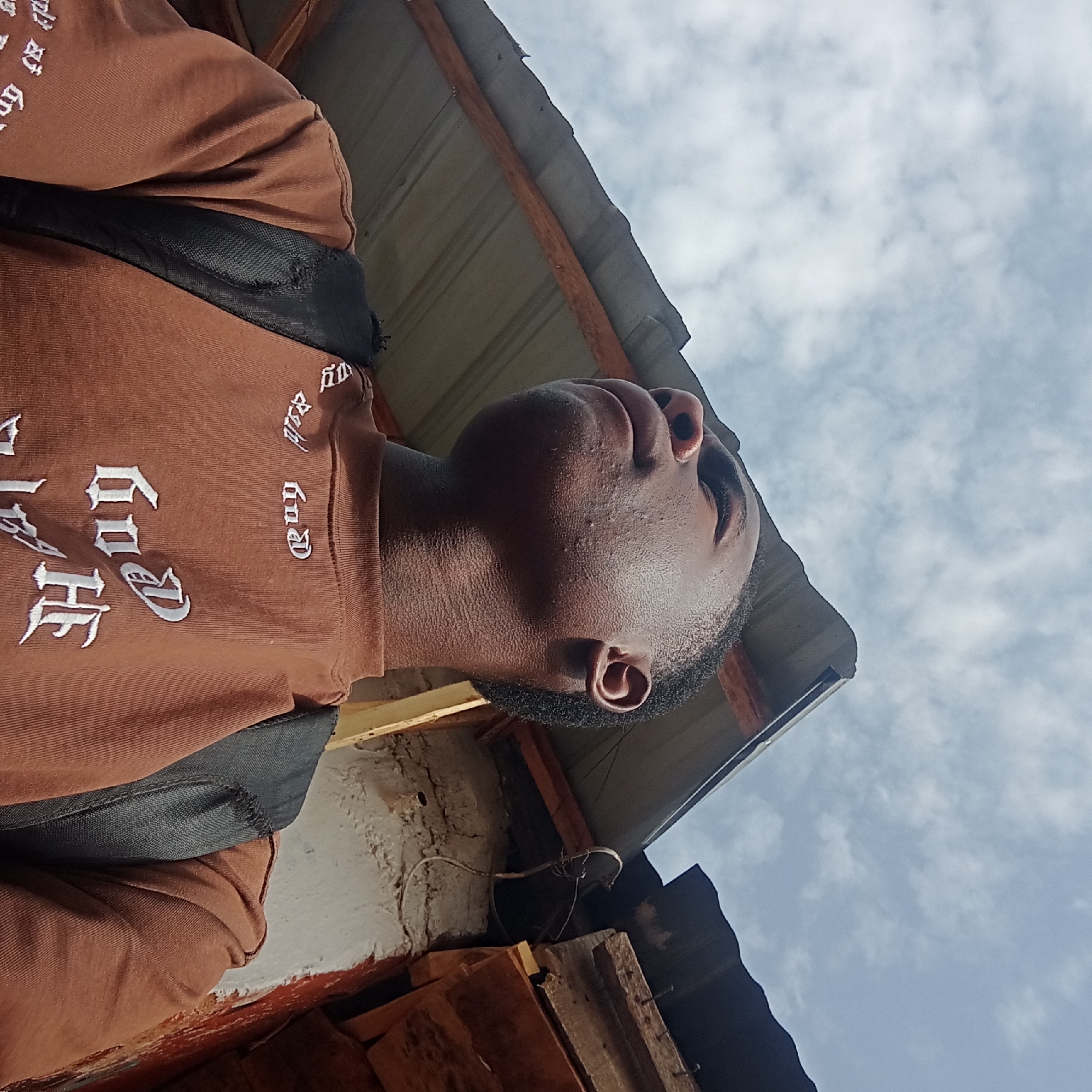 A low-angle shot captures a young Black man looking upwards and slightly to his right, with a bright, cloudy sky in the background. He wears a brown long-sleeved shirt adorned with white text in a gothic font, clearly displaying the words "THE," "GOD," and "and." A black strap, likely from a backpack, is visible across his left shoulder. His face has light stubble, and his left ear is visible.
The setting appears to be outdoors during daytime. A corrugated metal roof, supported by wooden beams, spans the upper left and central parts of the image, suggesting he is standing near or under a structure. Below the roof, a textured wall, possibly made of concrete or mud, shows patches of white and reddish paint or plaster, with some wires attached. Rough wooden planks are also visible towards the bottom of the frame. The man's expression is neutral as he gazes skyward. The environment cues align with a rustic or informal setting, consistent with the provided location context of Jos, Nigeria.
