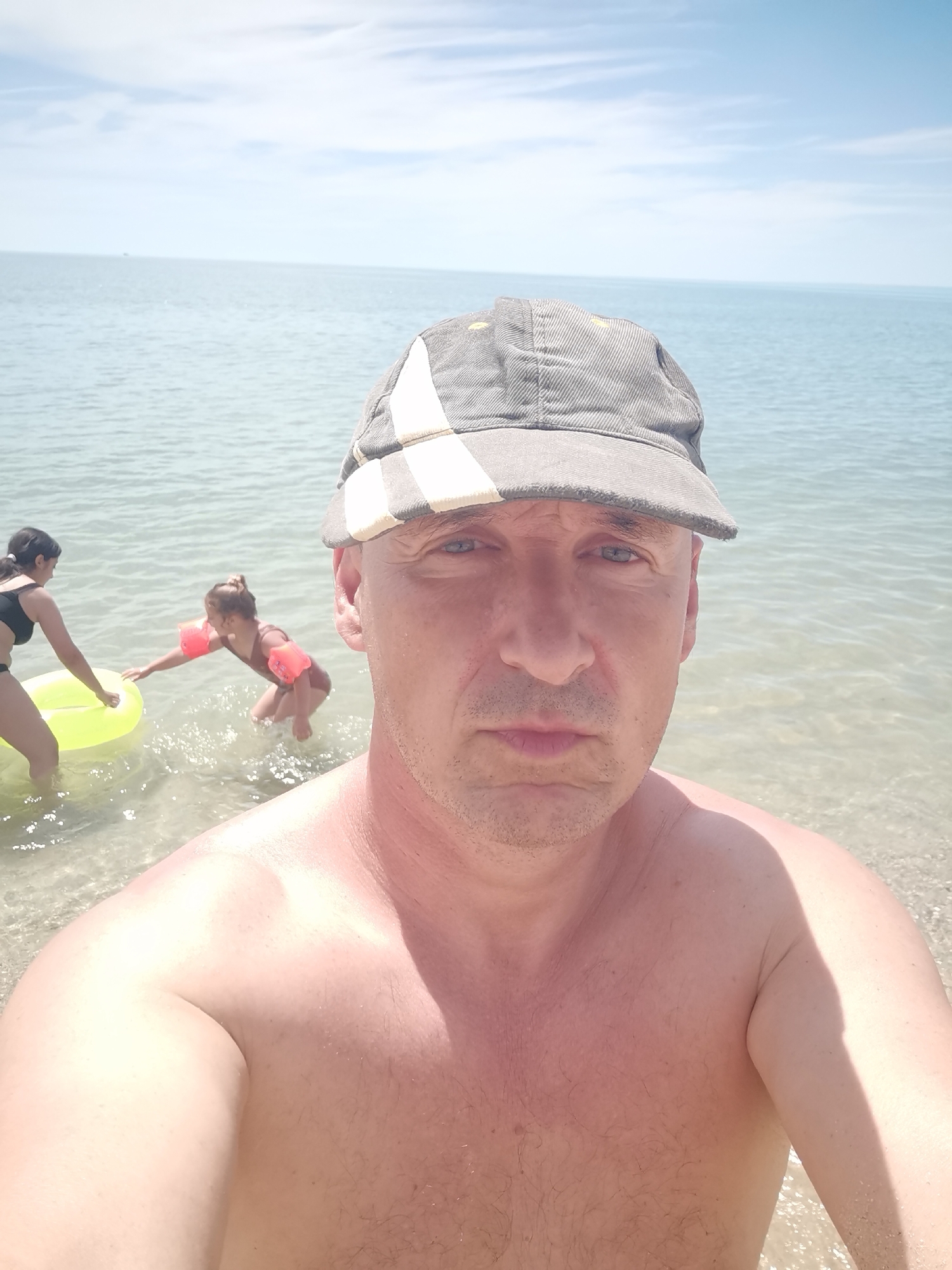 The image is a selfie taken by a man on a beach in Kranevo, Bulgaria. He is wearing a cap and is shirtless, with the sea and sky in the background. In the shallow water behind him, two children are playing. One child, wearing an orange swim vest, is reaching for a yellow inflatable ring held by another child. The water is clear and calm, and the sky is partly cloudy, suggesting it is a bright, sunny day. The man's facial expression is neutral, and he appears to be looking directly at the camera. The overall scene conveys a sense of a relaxed family outing at the beach.