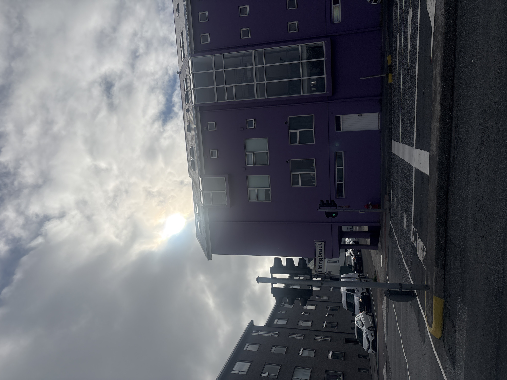 The image shows a street scene in Reykjavik, Iceland, with a prominent purple building on the right. The street is paved with asphalt and marked with white lines indicating lanes and pedestrian crossings. A traffic light is visible, with the green light illuminated, suggesting traffic is permitted to proceed. A street sign reads "Hringbraut," identifying the location. Several vehicles, including a white van and cars, are present on the street. The purple building features multiple windows and a large glass-paneled section. Adjacent to it, a gray building with many windows is also visible. The sky is overcast with patches of sunlight breaking through the clouds. The overall scene depicts a typical urban environment in Reykjavik.