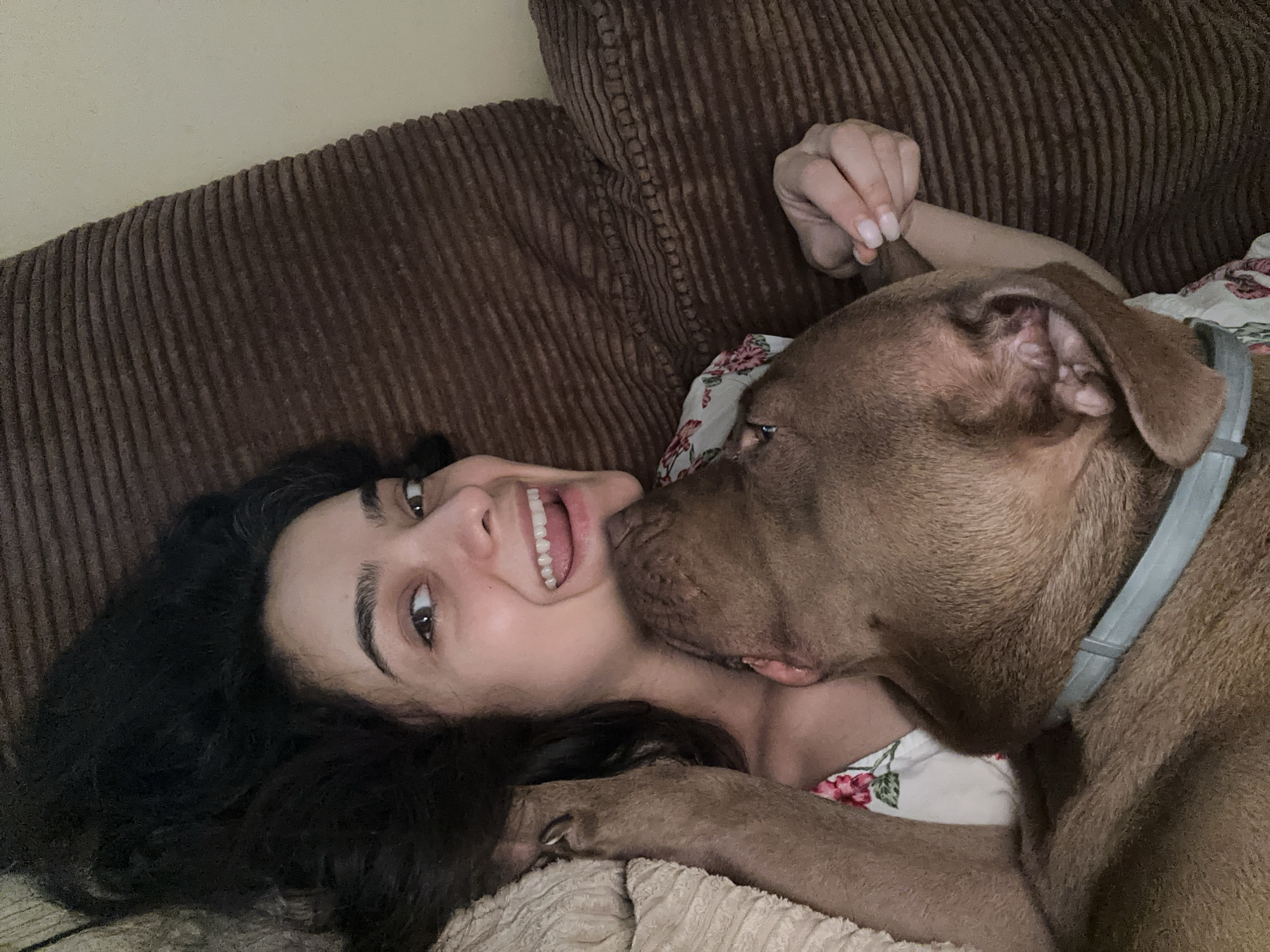 A young woman with long, dark curly hair lies on a brown corduroy couch, her head tilted back and her eyes wide open. A brown dog, possibly a pit bull mix, is leaning in and licking her cheek. The woman is smiling broadly, her mouth open and teeth showing. She is wearing a white top with a floral pattern. The dog has a light blue collar around its neck. The scene appears to be indoors, likely in a living room. The lighting is soft and warm, suggesting it might be late afternoon or evening. The woman's expression conveys happiness and affection, and the dog's action suggests a playful or affectionate interaction. There is no visible text in the image.