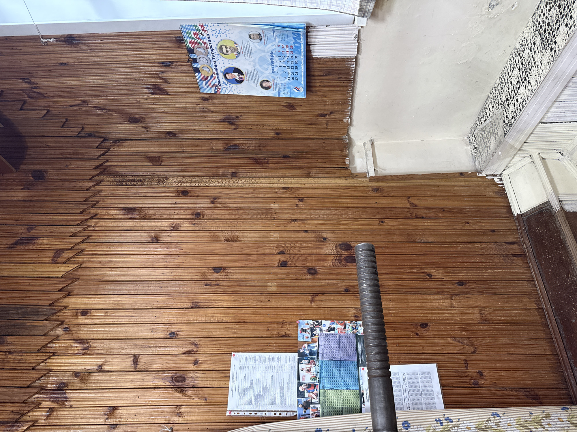 This is a media file showing an interior scene with wooden paneling on the wall and ceiling. In the upper portion of the image, a calendar for October 2014 hangs on the wall. The calendar features images of athletes and Olympic rings, with the text "Олимпийцы Татарстана" (Olympians of Tatarstan) prominently displayed. Below the calendar, there are some white, corrugated panels.
In the lower portion of the image, a collection of papers and printed materials is visible on the wooden wall. Among these papers are what appear to be charts or schedules, with some areas colored purple, blue, and green. A tall, cylindrical metal object with ribbed texturing stands vertically, partially obscuring some of the papers. The overall scene suggests a room that might be used for display or storage, possibly in a sports-related context given the calendar's theme. The lighting suggests an indoor environment, likely during the daytime. There are no strong indicators of weather conditions. The presence of the calendar and papers implies a specific time and potentially a commemorative or informational purpose.