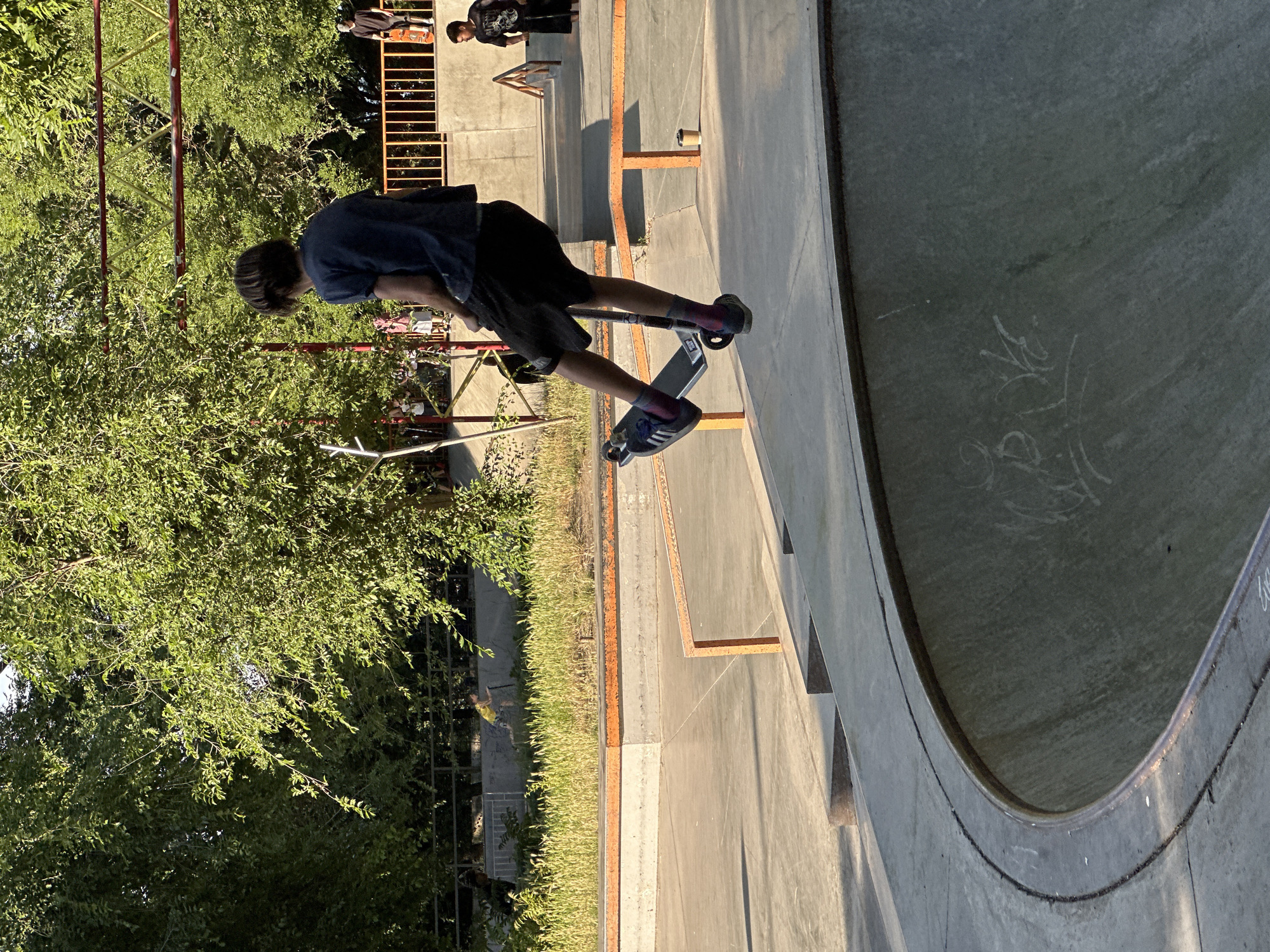 A person is on a scooter in a skatepark. The person is in mid-air, with their legs bent and the scooter positioned below them. The person is wearing a dark t-shirt, black shorts, and colorful socks. They are in the foreground of the image, with a concrete skatepark in the background. There are trees and a fence in the background, suggesting an outdoor setting. The lighting indicates it is daytime, likely late afternoon due to the warm tones. Other people and skatepark features are visible in the background.