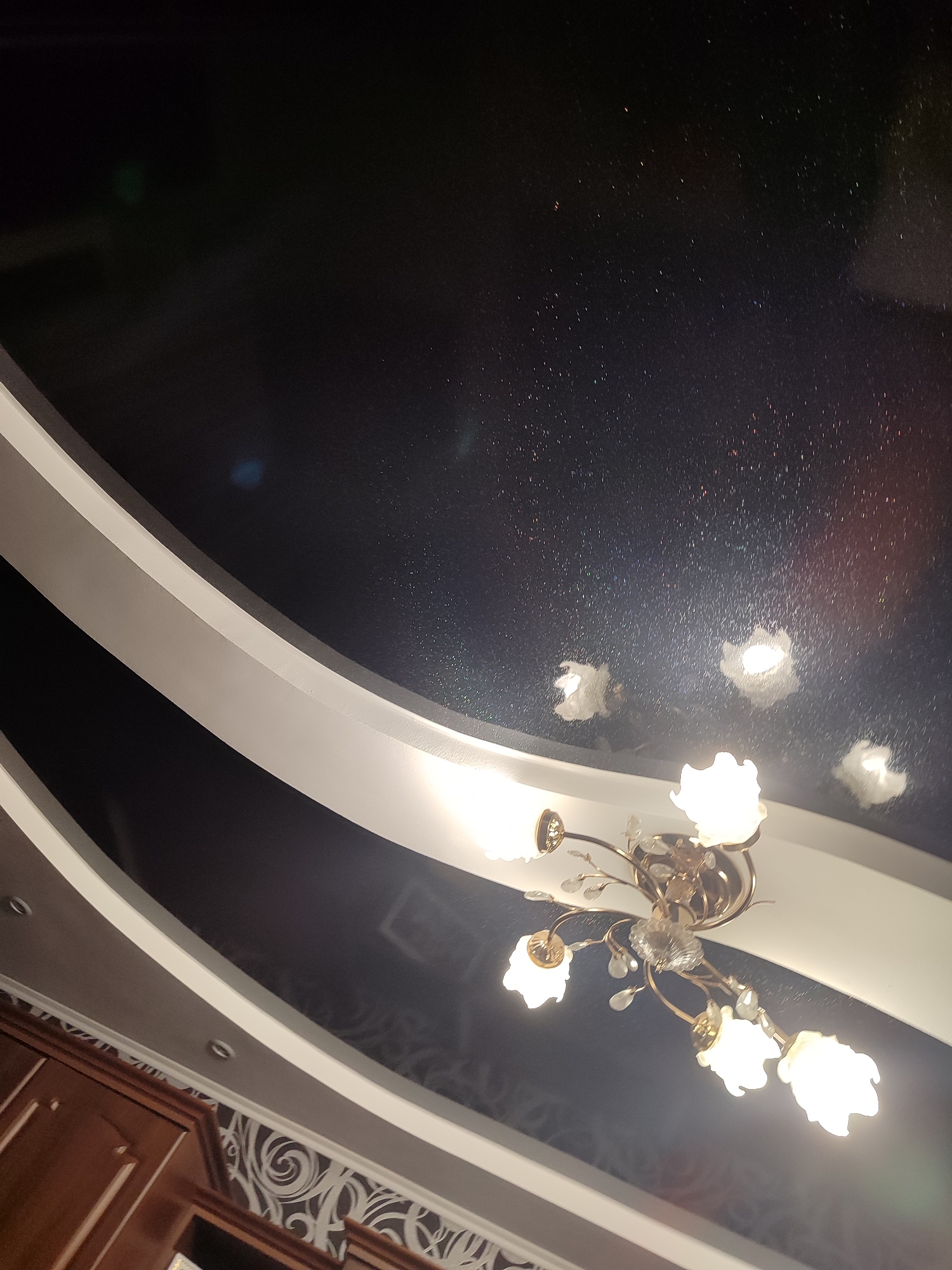The image shows an interior scene, likely a living room or dining room, with a ceiling featuring a dark, possibly black, stretch ceiling with a starry effect. A modern chandelier with multiple white, flower-shaped lights and decorative crystal accents is mounted centrally. The ceiling has curved white decorative elements. Below the ceiling, a portion of wooden cabinetry and wallpaper with a swirling pattern are visible. The image is taken from a low angle, looking upwards. There is no indication of the city of Mykolaiv, Ukraine, or any specific location. The scene depicts a residential interior, focusing on the ceiling and light fixture.