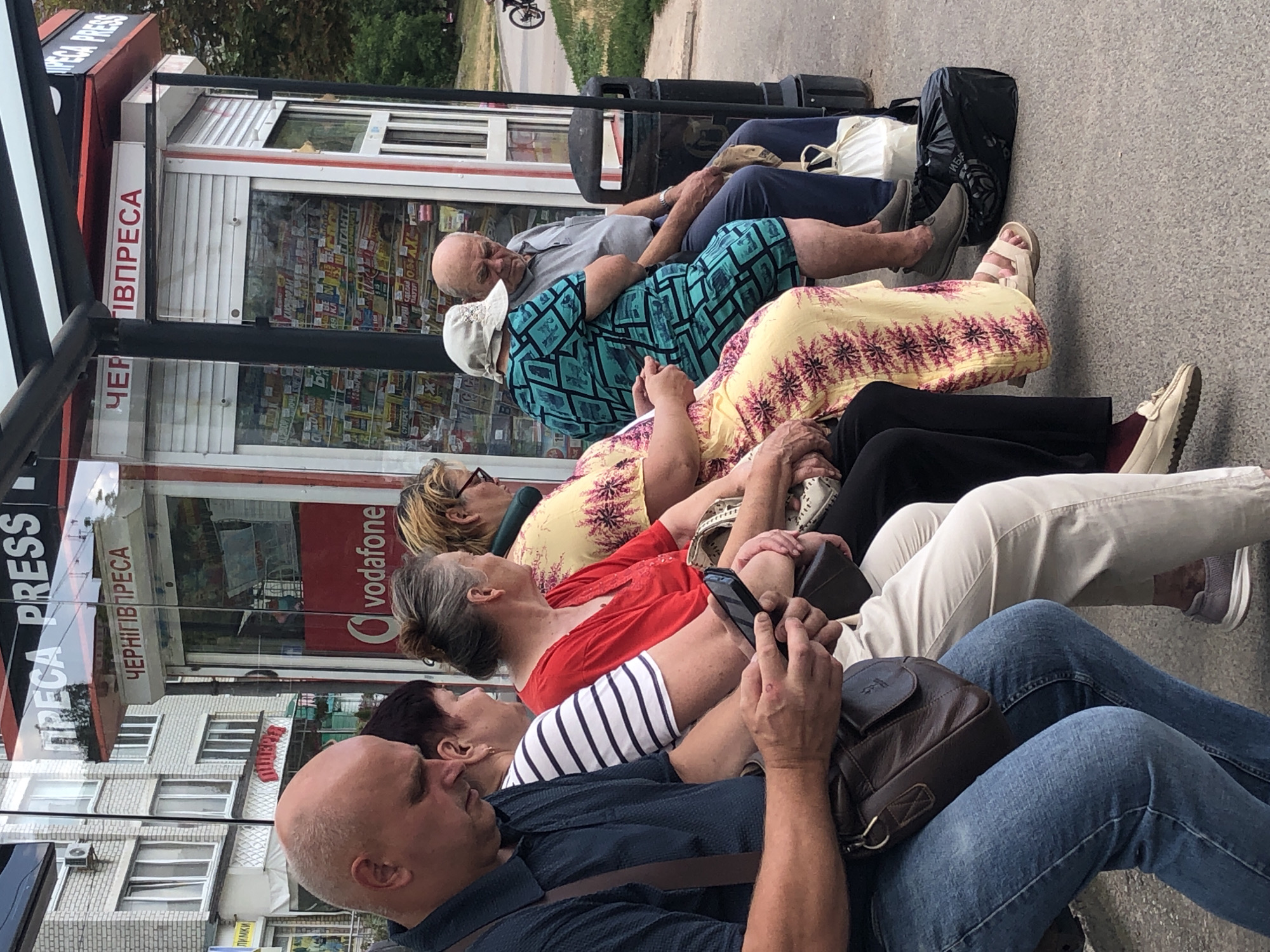 The image depicts a group of five individuals, likely waiting at a bus stop or similar public transit waiting area, in Chernihiv, Ukraine. The setting appears to be outdoors, with a covered structure overhead suggesting a bus stop shelter. The structure features signage with the words "ПРЕСА PRESS" and "ЧЕРНІГІВП РЕСА," indicating a press kiosk or newsstand. Several people are seated on a bench, facing away from the camera, with varying levels of engagement.
In the foreground, a bald man wearing a dark blue polo shirt and jeans is looking down at his mobile phone, holding it in his left hand. To his right, a woman with dark hair wearing a striped shirt is seated next to him. Further to the right, a woman with gray hair is wearing a bright red top and is looking towards her right. Next to her, a woman in a yellow dress with a floral pattern is also looking to her right. The furthest person on the right is an elderly man in a gray shirt and dark pants, also looking down.
Various objects are visible, including a dark brown leather bag on the lap of the man in the foreground, a handbag next to the woman in the red top, and a black trash bin with a white plastic bag on top next to the elderly man. In the background, a newsstand is stocked with magazines and newspapers. A building with windows and an air conditioning unit is visible behind the newsstand, suggesting an urban environment. The pavement is visible in the foreground and the ground extends into a road with a bicycle visible in the distance. The overall atmosphere suggests a moment of quiet waiting in a public space.