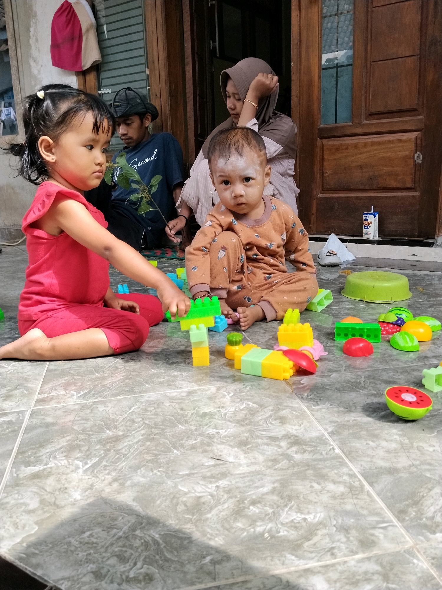 The image depicts a domestic scene with four individuals. In the foreground, a young girl in a red outfit is playing with colorful plastic building blocks on a tiled floor. Next to her, a baby in an orange outfit is also seated on the floor, looking towards the camera. Behind them, a man wearing a dark t-shirt and hat is looking down, and a woman in a hijab is leaning her head on her hand. The background shows a wooden door and part of a building's facade. The scene is likely set in an outdoor or semi-outdoor area, possibly a porch or yard, given the tiled flooring and the presence of building elements. A carton of milk with a straw is visible on the ground near the door. The environment suggests a residential setting in Panggungan, Indonesia.