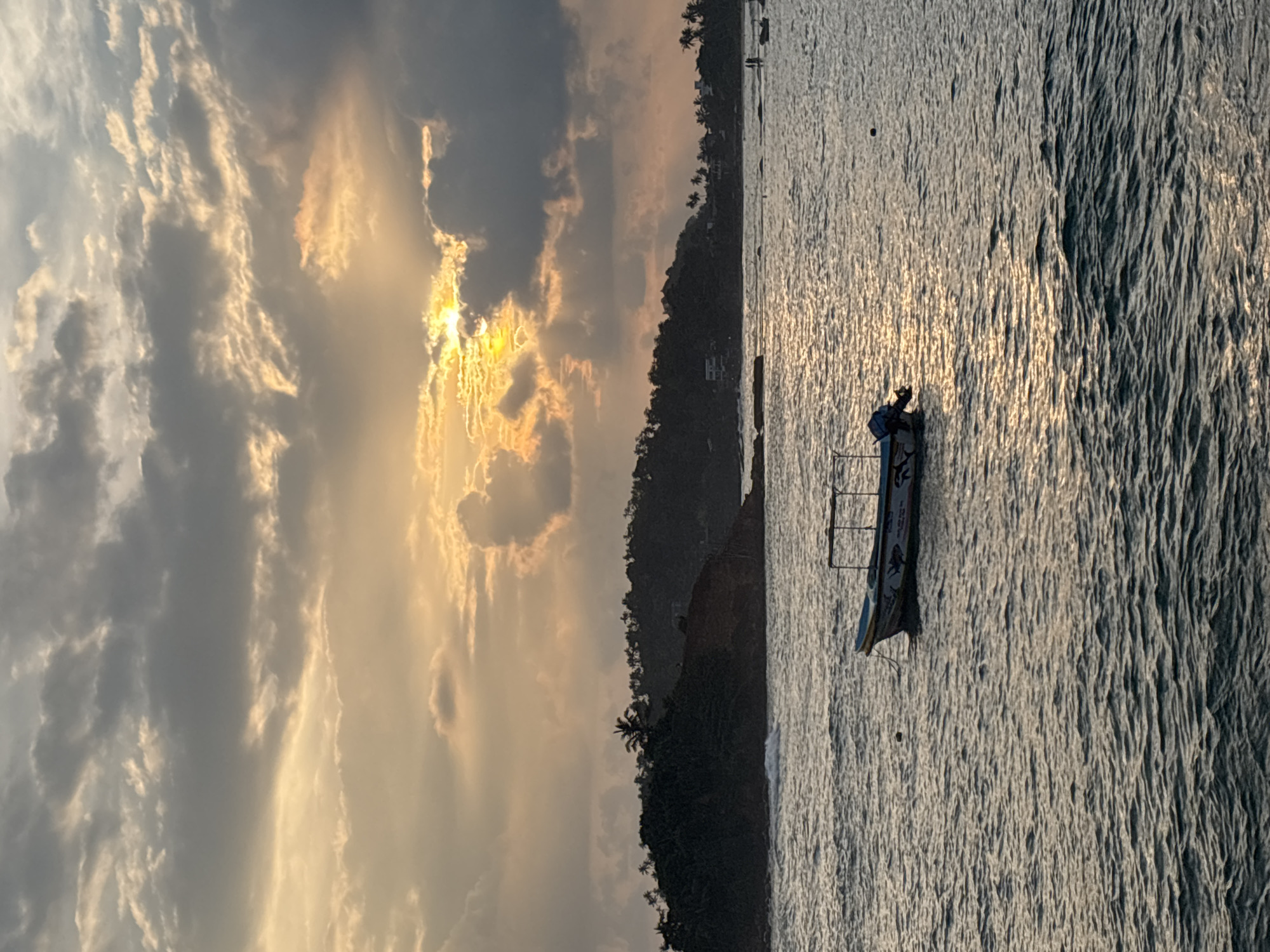 The image depicts a coastal scene in Thalaramba, Sri Lanka. The sky is dominated by grey and gold clouds, with the sun obscured behind them, creating a dramatic lighting effect. Below the clouds is the coastline, densely covered in trees. The ocean occupies the right portion of the image, its surface reflecting the sunlight. A small fishing boat with a metal frame sits on the water's surface.