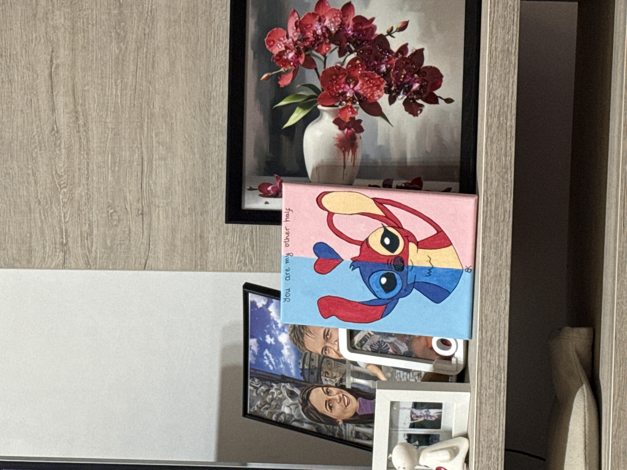 The image shows a cluttered shelf inside a wooden cabinet. On the top shelf, there is a framed painting of red flowers in a white vase. Below it is a canvas with a cartoon drawing of Stitch, with the text "You are my other half" written vertically along the left side. The bottom shelf contains a framed photograph of a smiling couple, and in front of it is a white picture frame with small objects inside. There is no indication that the location is in the city of Plovdiv, Bulgaria.