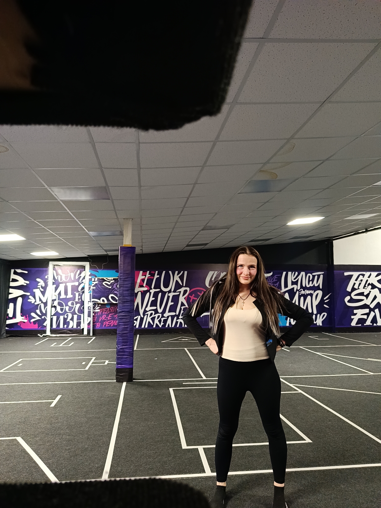 A young woman with long, dark hair stands confidently in an indoor facility, facing the camera with a slight smile. She is wearing a light beige or cream-colored fitted top, a black open jacket, black leggings, and dark socks or low-cut shoes. Her hands are on her hips, and she stands centered in the frame.
The setting appears to be a recreational or sports area, possibly a specialized gym. The floor is dark grey with numerous white lines marking out multiple courts or activity zones, similar to those found in sports halls. A prominent purple padded pole, resembling a net support, stands vertically in the mid-ground.
The walls are decorated with vibrant purple murals featuring white and some pink graffiti-style lettering. Visible text on the left wall includes Russian words such as "МИР" (world) and "ЖИЗНЬ" (life). In the center, English text reads "FEELOKI" (or similar stylized word) and "NEVER SURRENDER." Further right, more Russian text, "ЦЕЛЫЙ МИР" (whole world) and "ЭТОТ" (this), can be seen, along with what appears to be "FIFTH SIX".
The ceiling is made of white acoustic tiles with visible grid lines and several recessed rectangular fluorescent light fixtures illuminating the space. The overall atmosphere suggests an active, modern indoor environment in Chaikovsky, Russia. There are no other people visible, and no specific activity is actively taking place; the woman is posing for the photo.