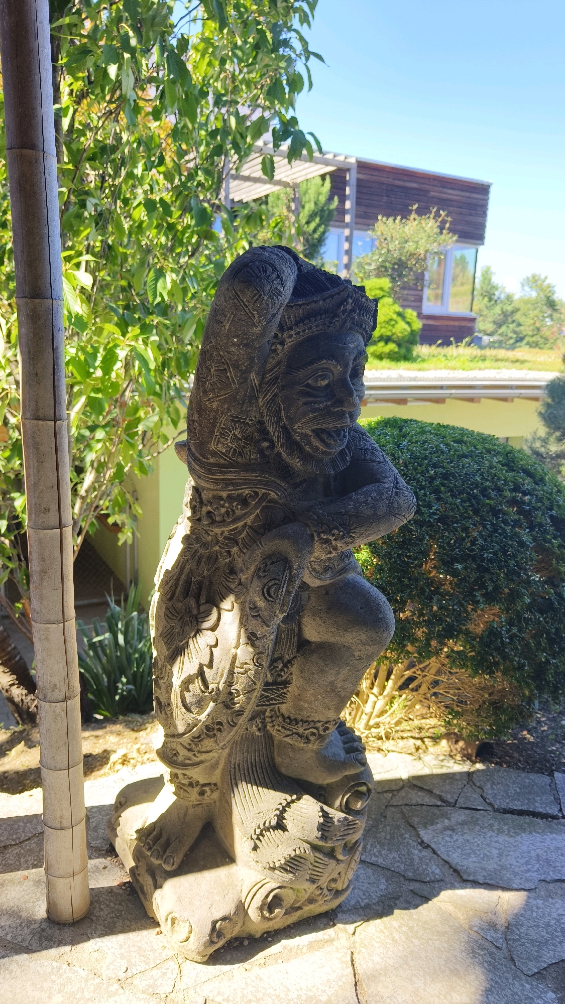 The image shows a weathered, stone statue of a mythological figure positioned on a cracked stone patio. The statue, dark gray in color, depicts a creature with an ornate headdress, a fierce expression with an open mouth, and detailed ornamentation on its clothing and body. It appears to be in a crouching or kneeling posture, with its arms folded across its chest. To the left of the statue, a segment of a bamboo pole, textured and lighter in color, stands vertically. Behind the statue, lush green foliage from a tree is visible, with sunlight casting dappled shadows. Further in the background, a modern building with wooden cladding and large windows is partially obscured by vegetation. A well-maintained, rounded green hedge is prominent in the mid-ground, to the right of the statue. The sky above is a clear, bright blue. The overall setting suggests an outdoor, possibly garden or courtyard area, within a residential or architectural context, likely in Langenlois, Austria, given the context of the prompt.