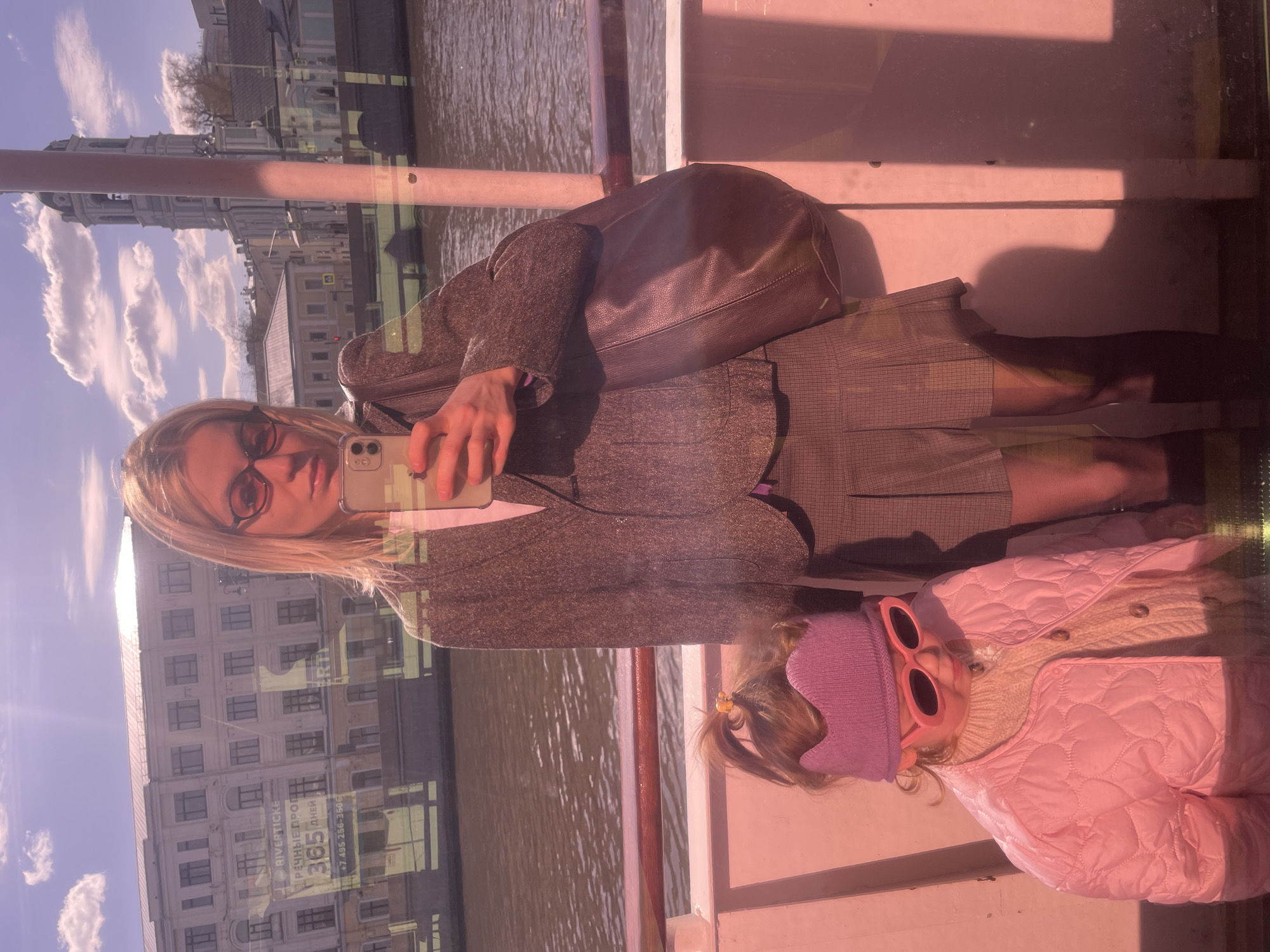 A woman and child are visible on a boat, as seen through a window. The woman is holding a smartphone, seemingly taking a selfie. The child is wearing sunglasses and a pink quilted jacket. The reflection in the glass shows a building in Moscow, Russia.