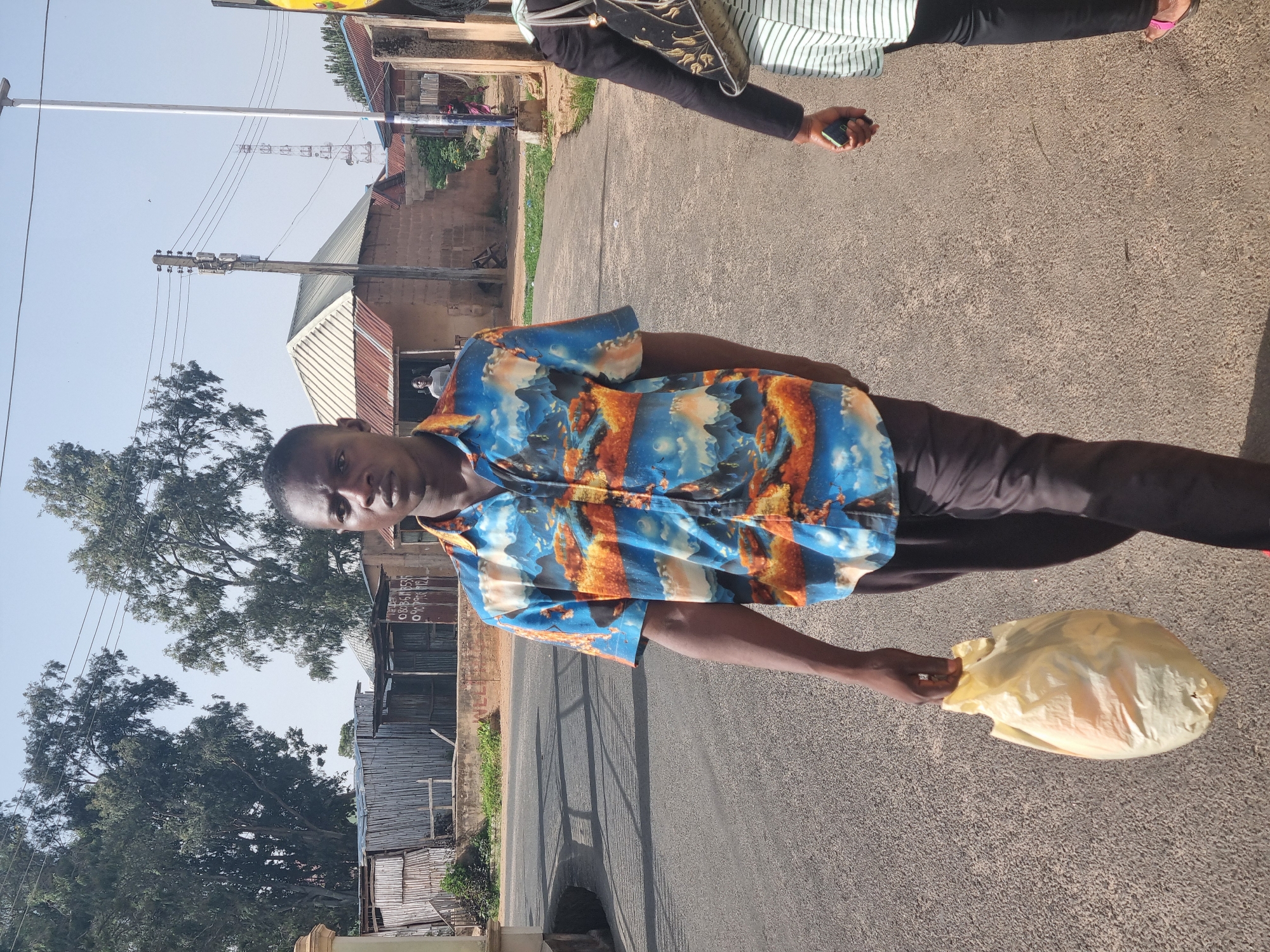 A man stands on a paved street in Jos, Nigeria, holding a plastic bag in his right hand. He wears a patterned blue and orange short-sleeved shirt and dark pants. The street is surrounded by buildings, including a corrugated metal structure, a more substantial brick building, and power lines overhead. A person wearing a striped shirt is partially visible in the upper-right corner. Shadows are cast on the ground, indicating strong sunlight.