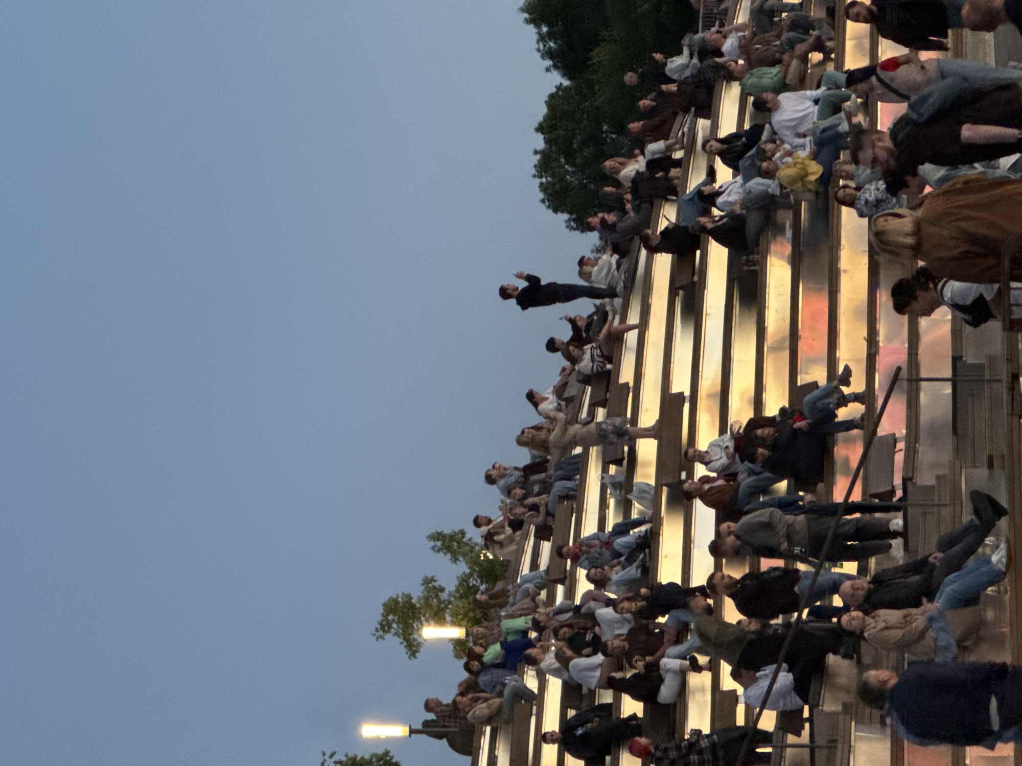 This image depicts an outdoor amphitheater filled with people attending an event. The seating is tiered, with individuals and small groups scattered across the steps. The lighting suggests it is dusk or early evening, with warm, artificial lights illuminating the lower tiers of the amphitheater. The sky is a clear, pale blue, indicative of twilight.
The amphitheater is characterized by its modern design, featuring wide, flat steps made of a material that appears to be wood or a composite, with metal accents or lighting elements integrated into the structure. Trees and foliage are visible in the background, suggesting a park or public space setting.
People are seen in various states of engagement, some sitting and conversing, others looking towards an unseen focal point, possibly a stage or performance area. The attire of the attendees is casual, consistent with an outdoor public gathering.
While the image itself does not provide explicit landmarks for precise location identification within Nizhny Novgorod, Russia, this type of open-air amphitheater is a feature in modern urban public spaces. Such venues are often found in parks, cultural centers, or riverfront developments. Without further context or specific visual cues linking it to a known Nizhny Novgorod location, its exact placement within the city cannot be determined from the image alone.