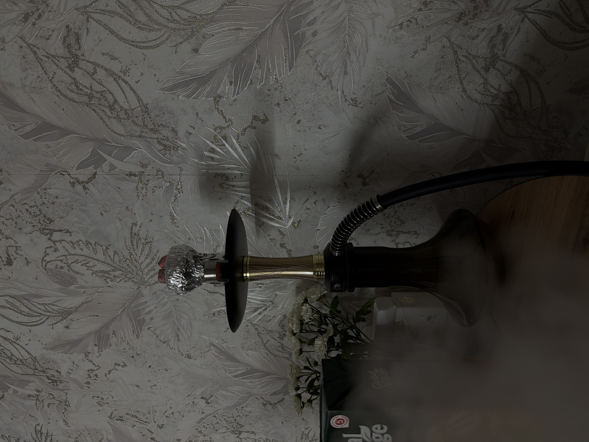 The image shows a hookah setup on a surface, likely a table. The hookah has a foil-covered bowl, a black hose, and a vase. The scene is indoors with a floral patterned wallpaper. A box with the words "Global Village" is visible next to the hookah. Based on the query, the location is inferred to be Reshetnikovo, Russia, although there is no independent confirmation of location in the image itself. There is a large amount of smoke coming from the hookah.