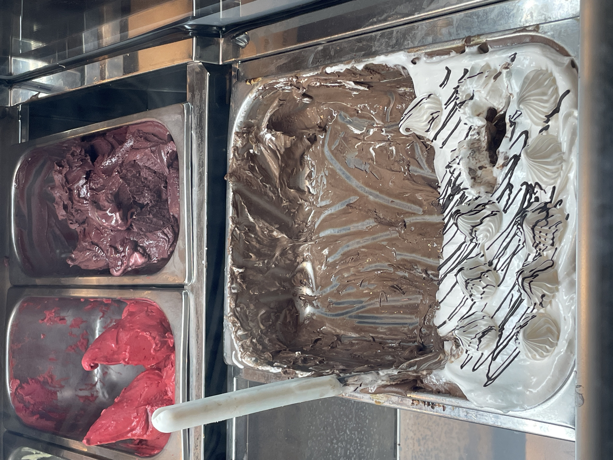 The image shows a display case of gelato in Buenos Aires, Argentina. Three rectangular stainless steel containers hold different flavors. The top left container holds a dark purple gelato. The bottom left container holds a bright red gelato. The large container on the right is divided into two sections. One section contains chocolate gelato with swirls of white. The other section contains white gelato decorated with swirls of chocolate and meringue. A white plastic ice cream scoop lies in the red gelato container. The display case is stainless steel. The lighting is bright and even.