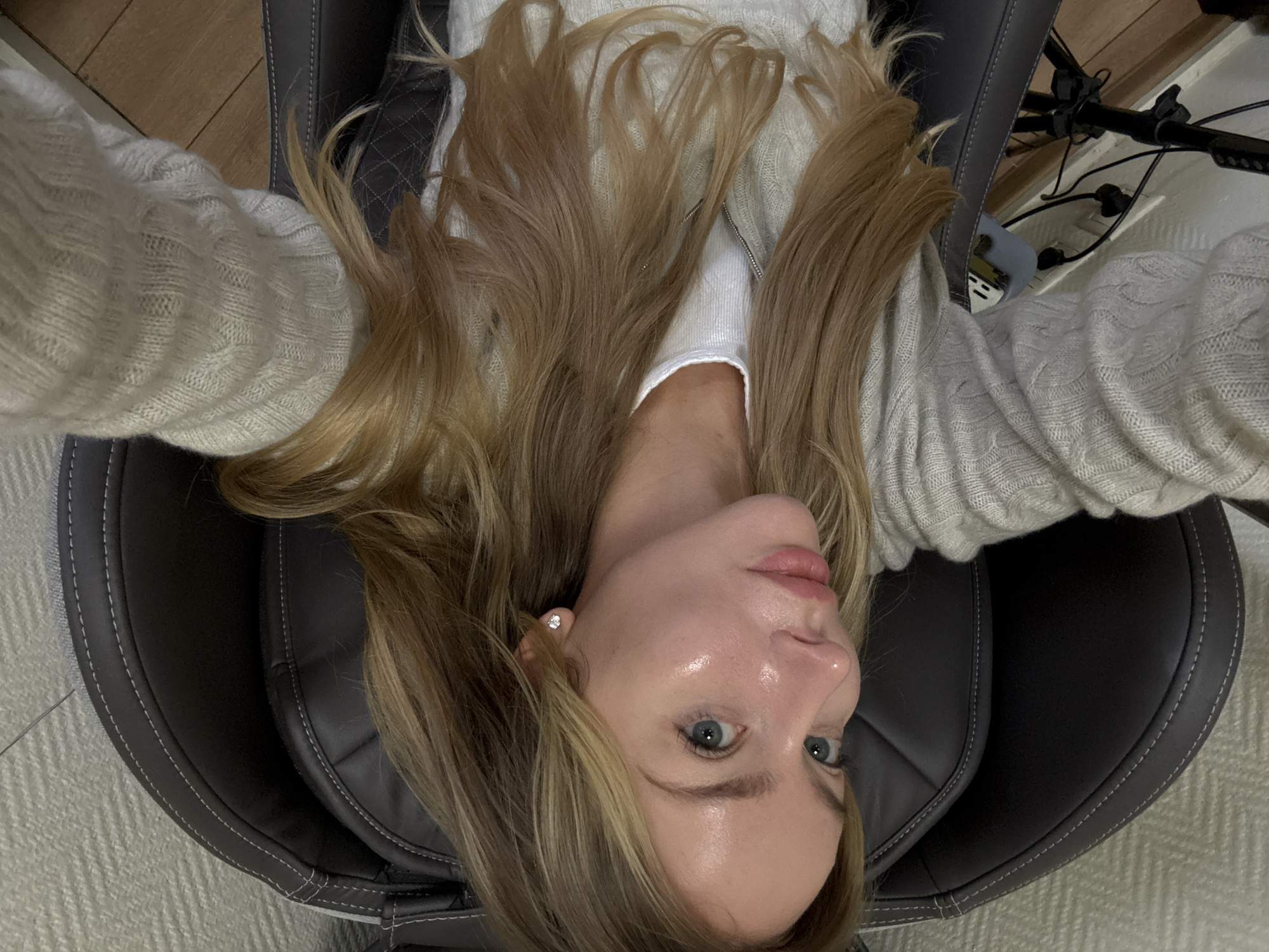 A woman with light skin, blonde hair, and blue eyes is lying in a gray chair. She is wearing a white shirt under a light gray sweater with a textured pattern. The chair is dark gray with light gray stitching and padding. In the background, there is a light-colored floor and some equipment or furniture legs with wires. There is nothing to indicate this image was taken in Moscow, Russia.