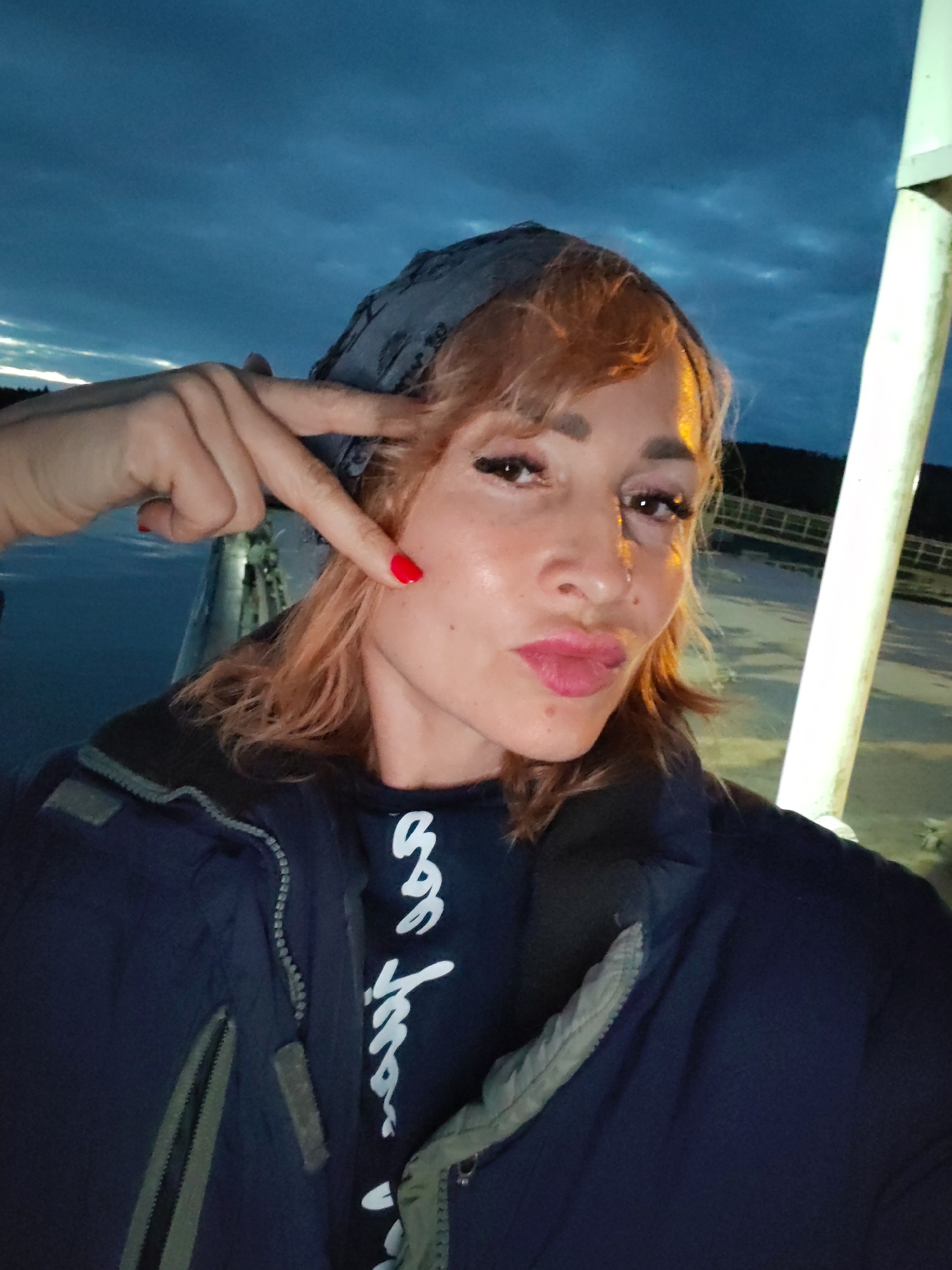 A woman with light brown hair and fair skin is pictured in a selfie. She is wearing a dark blue jacket over a black t-shirt with white writing. Her left hand is raised, with her index and middle fingers extended in a peace sign, pointing towards her cheek. She has red nail polish on her fingers and a mole on her left cheek. The background suggests an outdoor setting at dusk or night, with a cloudy sky and a lit structure to the right of the frame. The location is identified as Shcheleiki, Russia.