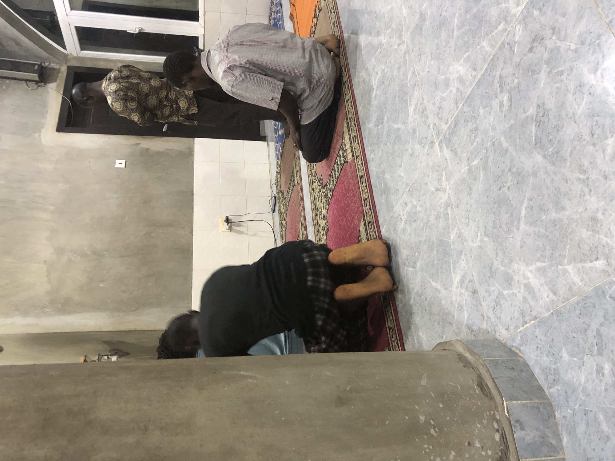 In a room with concrete walls and a marble-tiled floor, possibly in Ado, Nigeria, three men are engaged in prayer. Two of the men are kneeling on prayer mats. The man in the foreground on the right, wearing a checkered shirt and dark pants, is kneeling with his feet tucked beneath him. To his left, a man wearing a black shirt and patterned skirt is also kneeling, with his bare feet visible. In the background, a third man stands facing a dark wooden door, wearing a patterned shirt and dark pants. The room is lit, with electrical outlets and a window visible. The prayer mats are colorful and patterned.