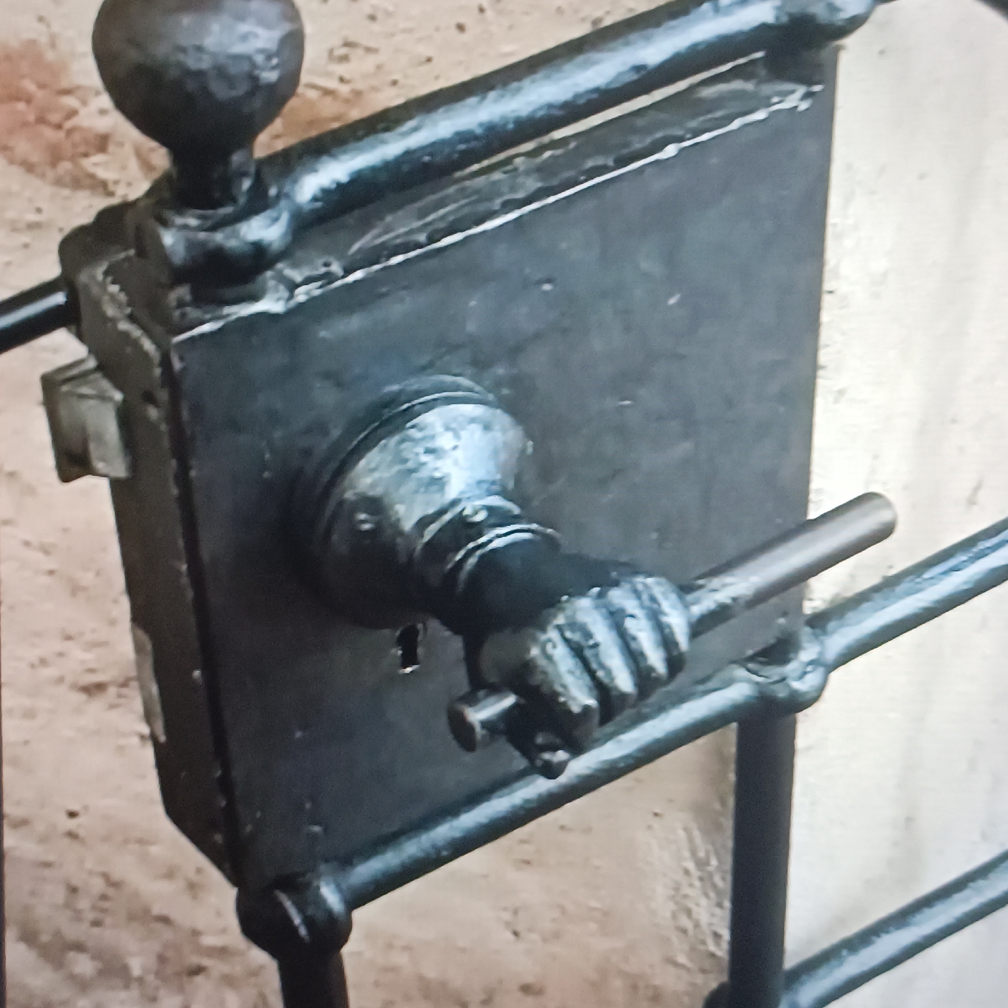 The image displays a close-up view of an ornate, black metal gate latch mechanism. The latch is robust and appears to be antique, featuring a decorative knob and a handle shaped like a clenched fist. The fist handle is gripping a horizontal bar, suggesting the latch is currently engaged or has just been operated. The latch assembly is attached to a larger, dark metal structure that is part of the gate, with vertical bars of the gate visible on either side. The background consists of a textured wall, likely brick or stone, and a lighter colored vertical surface, possibly another part of the building or wall. There is no indication of the specific location within Rüsselsheim am Main, Germany, as no identifying landmarks or text are visible. The lighting suggests an outdoor environment.