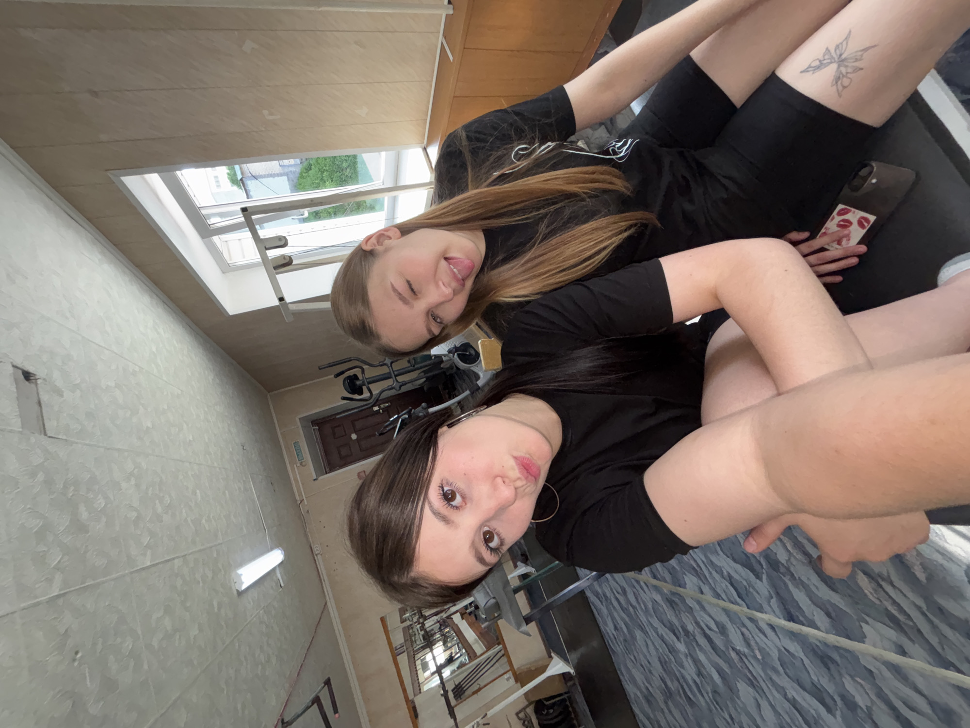 Two young women pose for a selfie inside what appears to be a gymnasium or fitness center. The woman closer to the camera has dark brown hair, brown eyes, and large hoop earrings. She is wearing a black t-shirt and has her arms crossed. The second woman has light brown hair and is winking at the camera, sticking out her tongue slightly. She is also wearing a black t-shirt and black shorts. On her thigh is a butterfly tattoo. She is holding a phone with a case decorated with lips.
The gymnasium contains exercise equipment. There is also a window with a view of greenery outside.
The image was most likely taken in Chaikovsky, Russia, because the location was specified in the request.