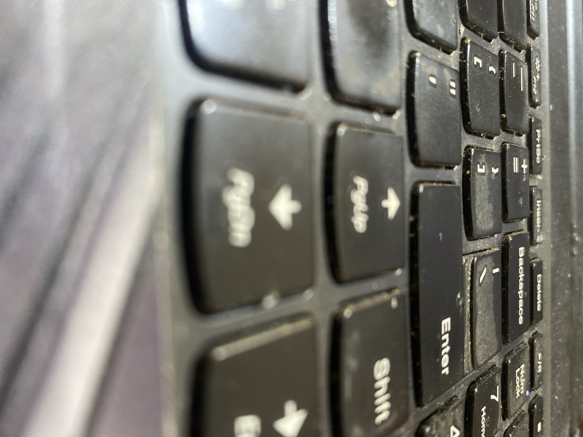 The image shows a close-up, low-angle view of a dirty black laptop keyboard. The keys are worn and covered in dust. Several keys are visible, including "PrtSc," "Insert," "Delete," "Backspace," "Enter," "Shift," "PgUp," and "PgDn." The focus is on the keys, with the background blurred. The scene appears to be indoors, likely a desk or workspace, but no specific location in North Jakarta, Indonesia, can be determined from the image alone.