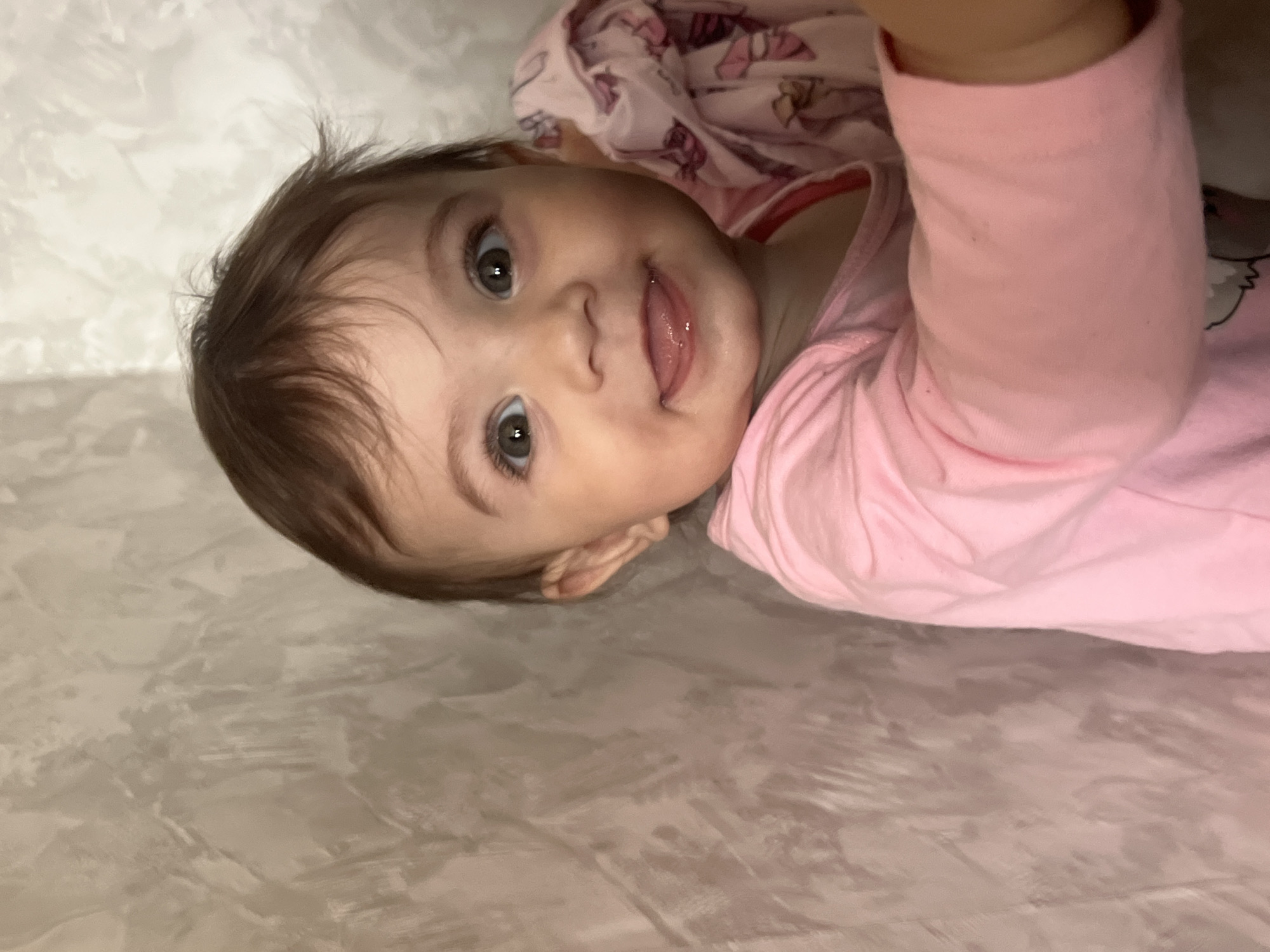 A baby with brown hair and hazel eyes, wearing a pink long-sleeved shirt and pink patterned clothing, is lying on a gray textured surface. The baby is looking at the camera with their tongue slightly protruding. No evidence suggests this photo was taken in Elena, Bulgaria.