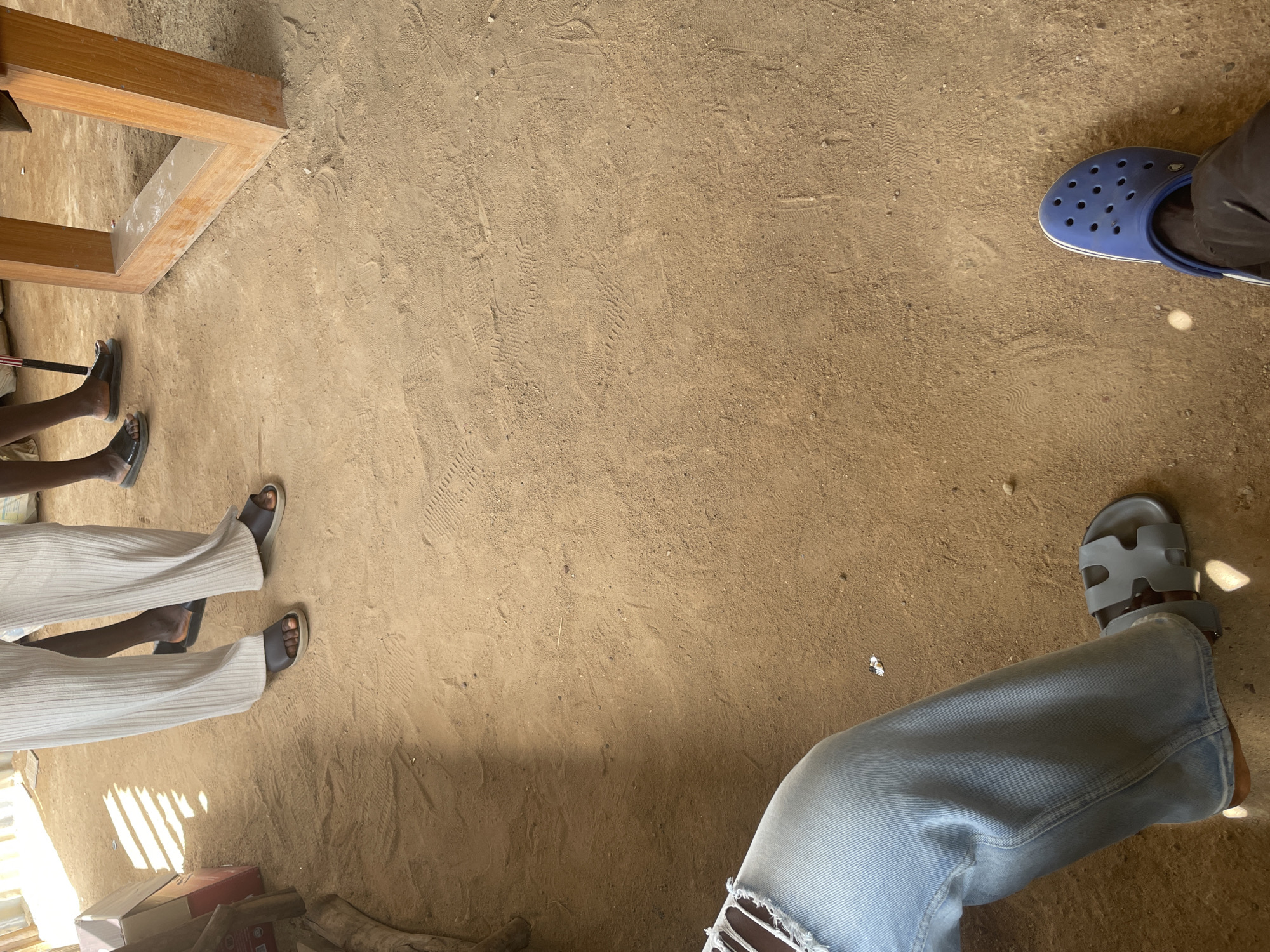 The image depicts a ground-level view of a sandy or dirt floor, likely an outdoor or semi-enclosed space. Several individuals' legs and feet are visible. On the left, a person is wearing ripped light blue jeans and light gray slip-on sandals. To their right, another person's legs are visible, clad in loose-fitting, light-colored pleated pants and dark slip-on sandals. Further to the right, another pair of dark slip-on sandals are visible, with one foot slightly obscured. In the lower right corner, a person is wearing dark pants and bright blue Crocs. The sandy ground shows numerous footprints and tire tracks. The environment suggests a rustic setting, possibly a market, workshop, or communal area. The lighting indicates it is daytime, with shadows cast across the floor. The specific location in the city of Durumi, Nigeria, cannot be determined from the visual information alone.
