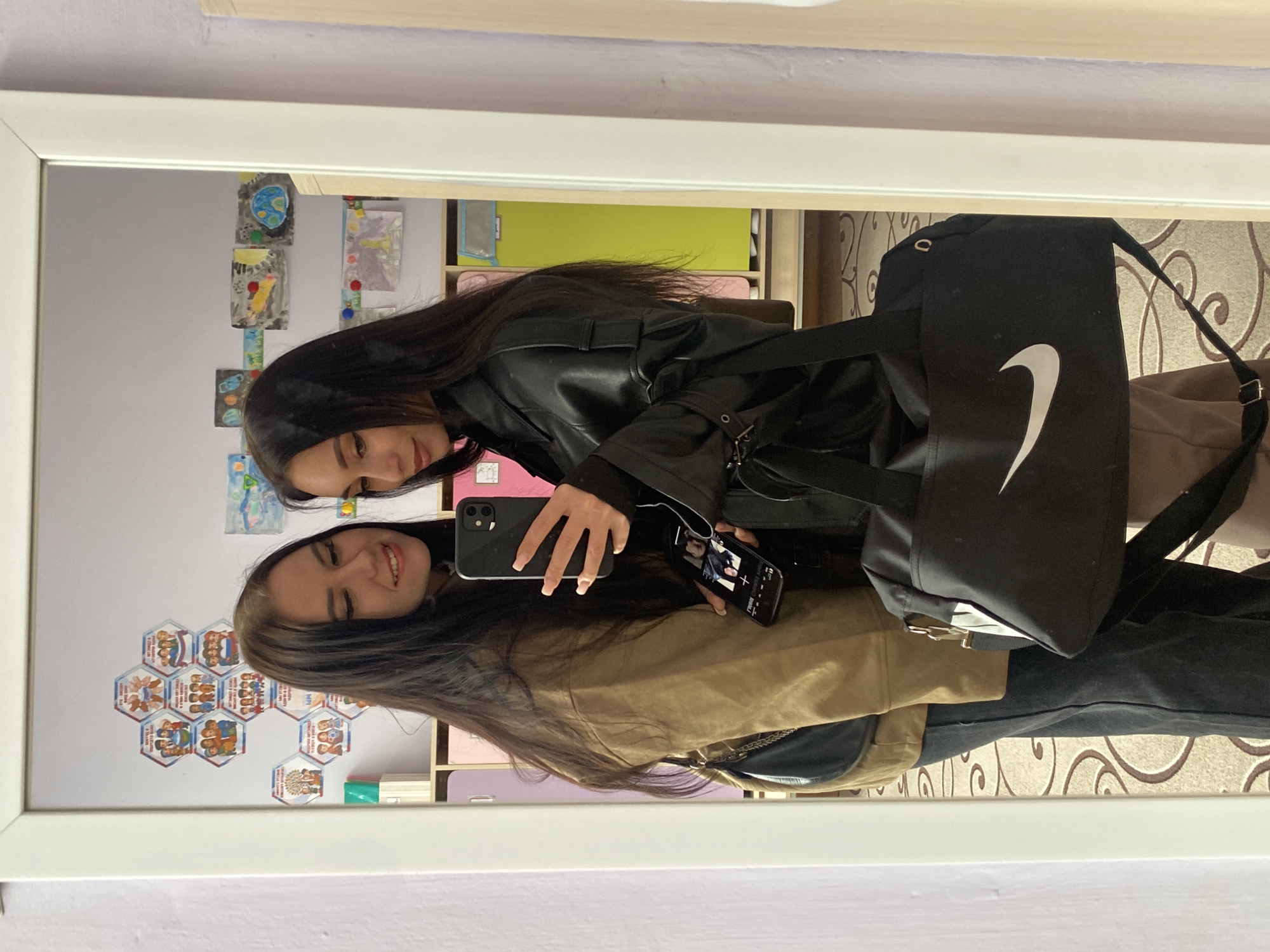 The image displays two young women reflected in a large mirror, posing for a photograph in an indoor setting. Both women have long, dark hair and are smiling, indicating a cheerful mood. The woman in the foreground wears a tan or light brown jacket and holds a black smartphone, which reflects her face. The other woman, dressed in a black leather-like jacket, also holds a smartphone, displaying an image on its screen. She carries a black Nike duffel bag featuring a prominent white swoosh logo, and a smaller black purse is visible near the first woman.
The background, visible through the mirror's reflection, features a wall decorated with several hexagonal awards or badges. These decorations include illustrations of people and text written in Cyrillic script, notably words like "ЧАЙКОВСКИЙ" (Chaikovsky) and "ОРЕЛ" (Orel), aligning with the specified location context of Chaikovsky, Russia. Above these, children's drawings, including one depicting a globe, are displayed. Faint reflections of shelves and other household items suggest a personal space, likely a home environment. A patterned carpet or rug is visible on the reflected floor. The scene appears to take place during the daytime.